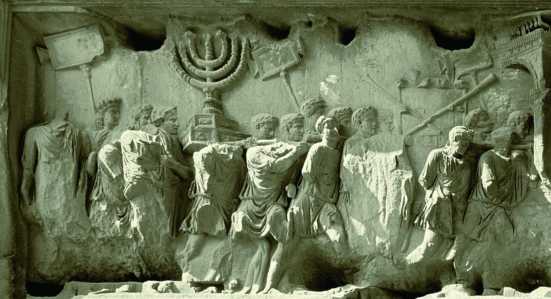 Escultura. Entalhe em uma pedra, representando um grupo de homens enfileirados, carregando um baú com diversos objetos, entre os quais um candelabro de sete braços, símbolo do judaísmo.