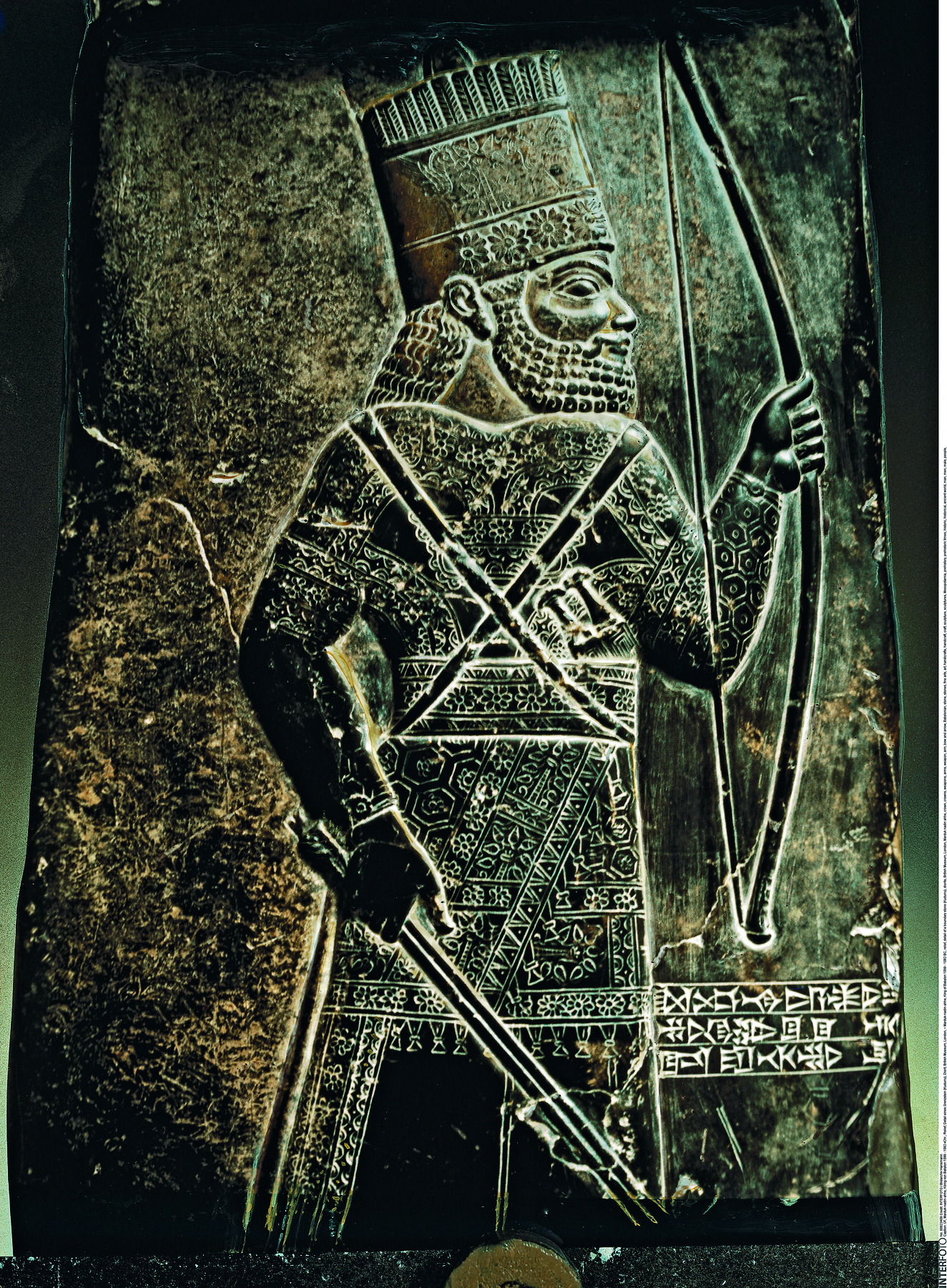 Baixo-relevo. Placa de pedra escura, com a imagem de um homem vestindo uma armadura e um chapéu comprido, visto de perfil, segurando um arco e uma flecha.
