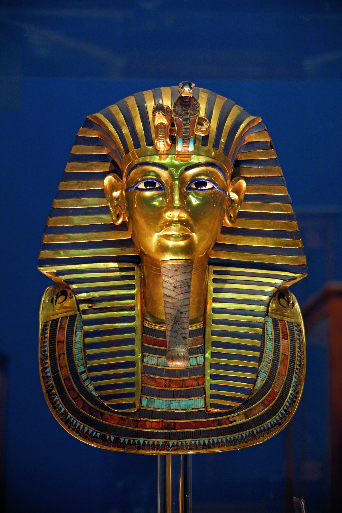 Máscara. Representação egípcia com traços humanos, dois olhos, um nariz e lábios, uma longa barbicha no queixo, e adornos ao redor da cabeça que caem sobre os ombros.