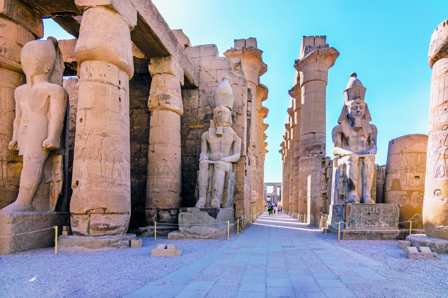 Fotografia. Vista das ruínas de um templo, com grossas colunas circulares e estátuas representando homens sentados sobre tronos.