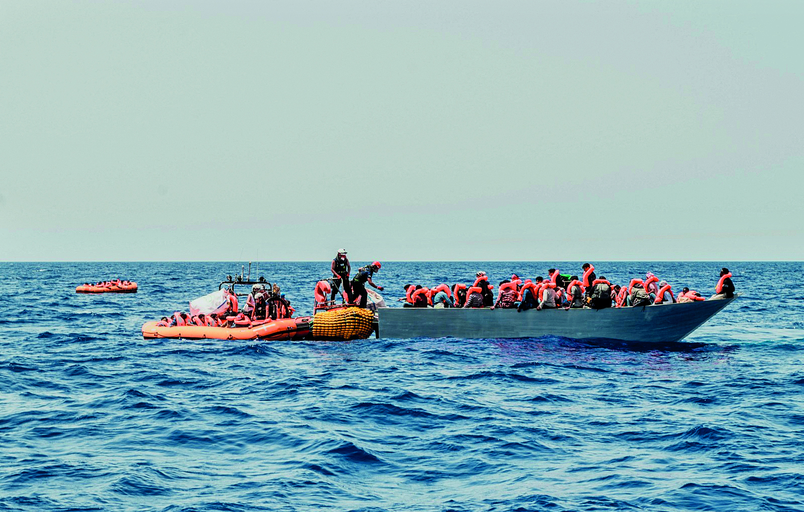 Fotografia. Pequenas embarcações repletas de passageiros sobre as águas do mar. Os passageiros vestem coletes salva-vidas laranjas.