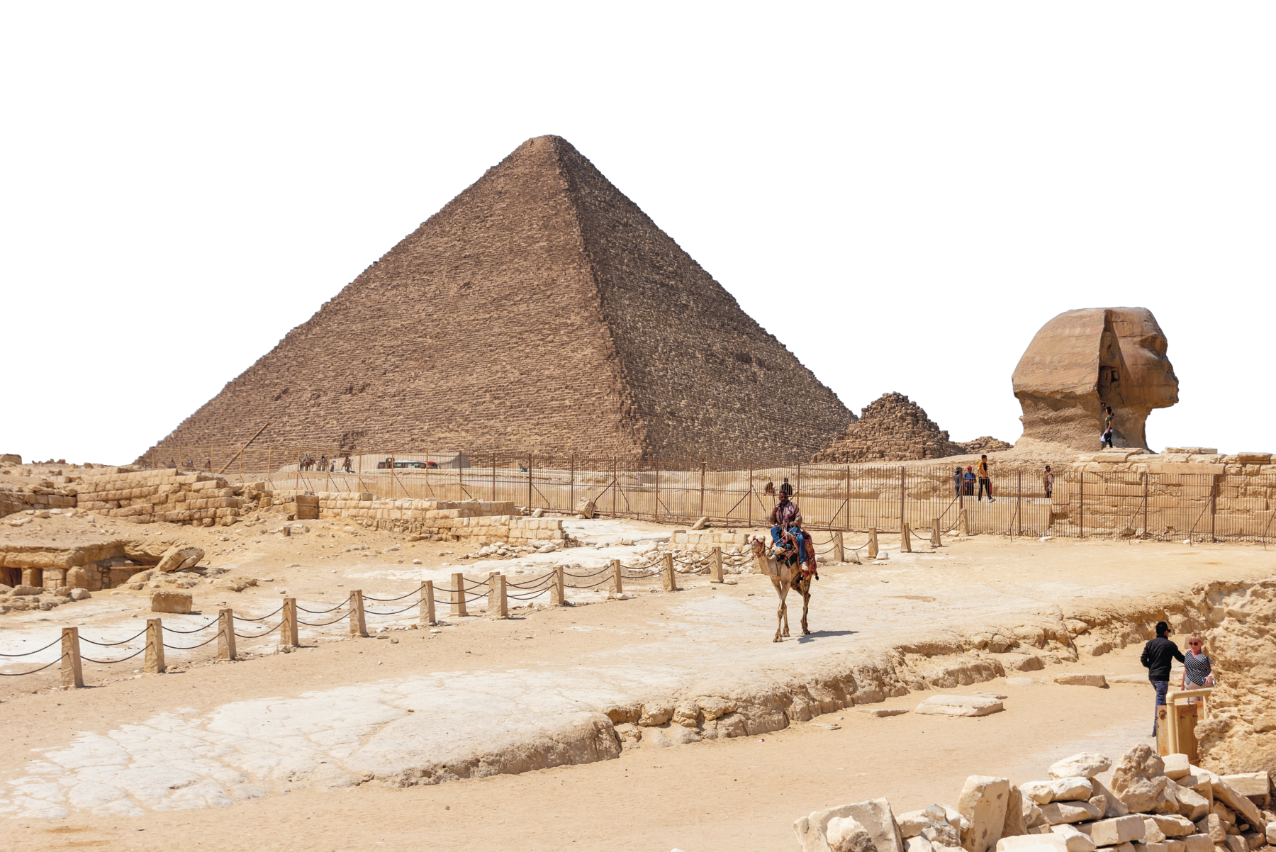 Fotografia. Vista de uma pirâmide de base quadrada e ápice triangular, e ao lado, uma esfinge em pedra, com cabeça humana. A área está cercada. Há um caminho pelo qual um homem sobre um camelo atravessa.