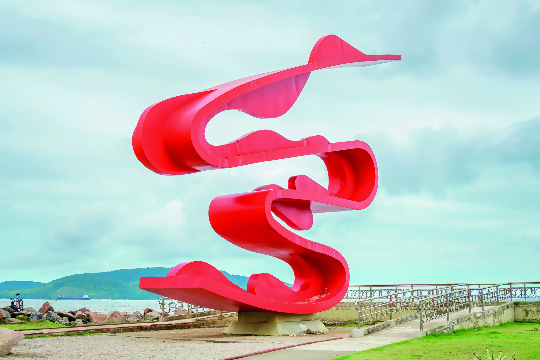 Fotografia. Destaque para escultura ao ar livre, com estruturas curvas, contínuas e assimétricas, na cor vermelha.
