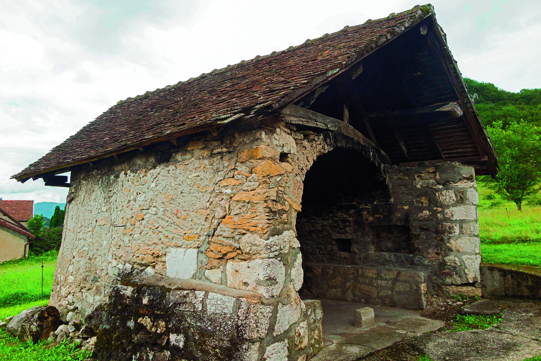 Fotografia. Imagem de uma construção com paredes de pedra, abertura central em forma de arco e telhado triangular.