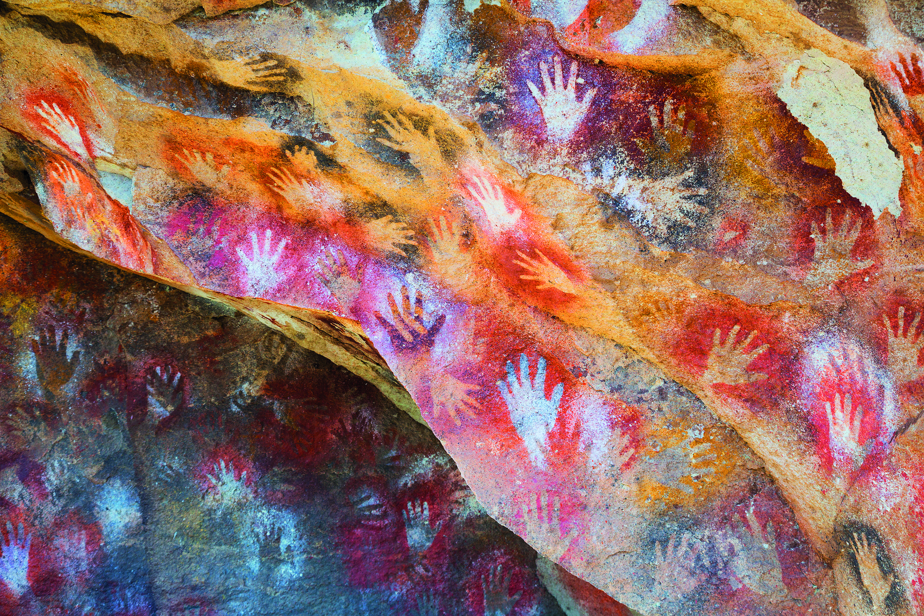 Fotografia. Contornos de diversas mãos, em tons de vermelho, amarelo, laranja, preto e branco, sobre as paredes de uma gruta.