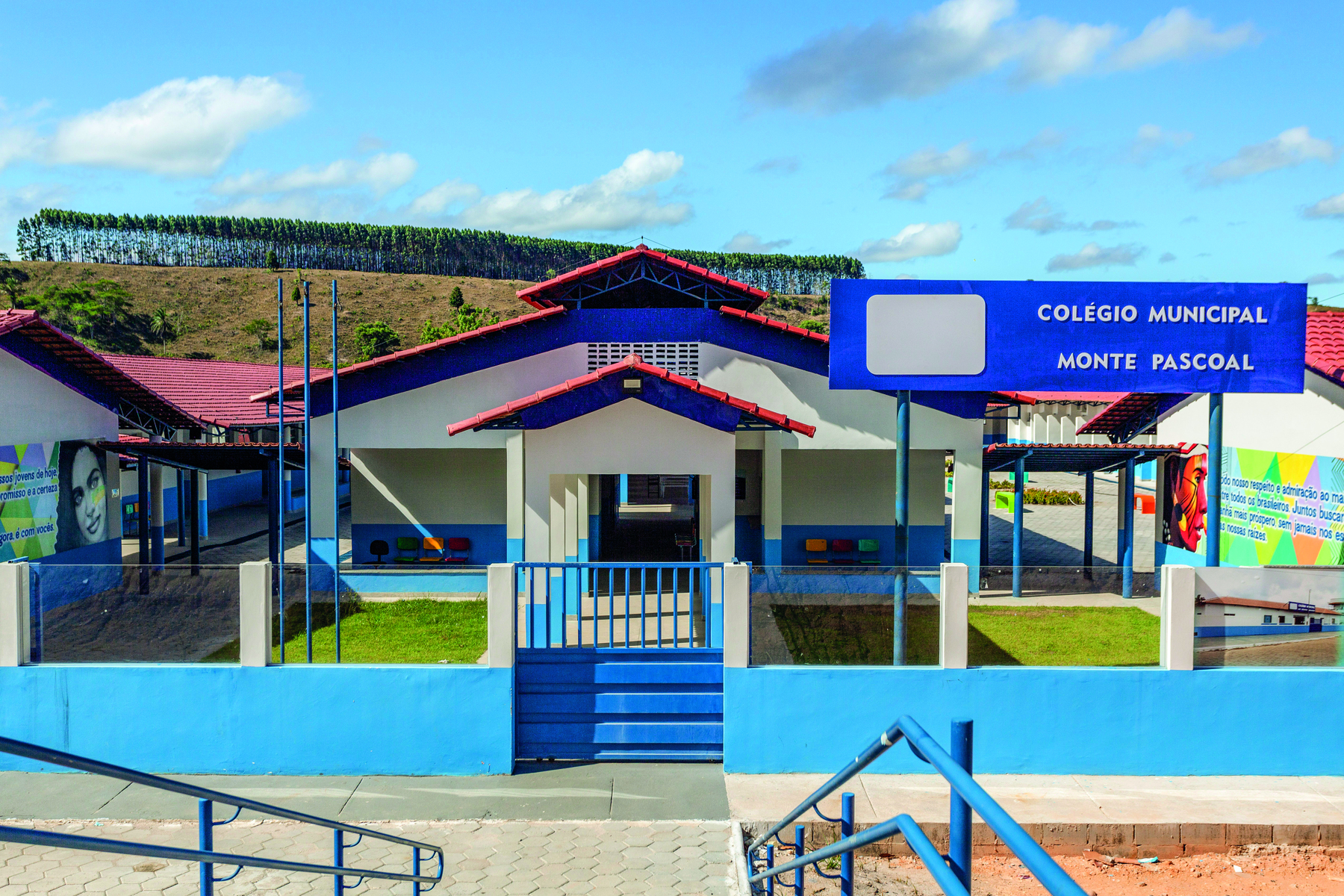 Fotografia. Fachada de uma escola, com paredes brancas e azuis, um portão central de entrada e área gramada.