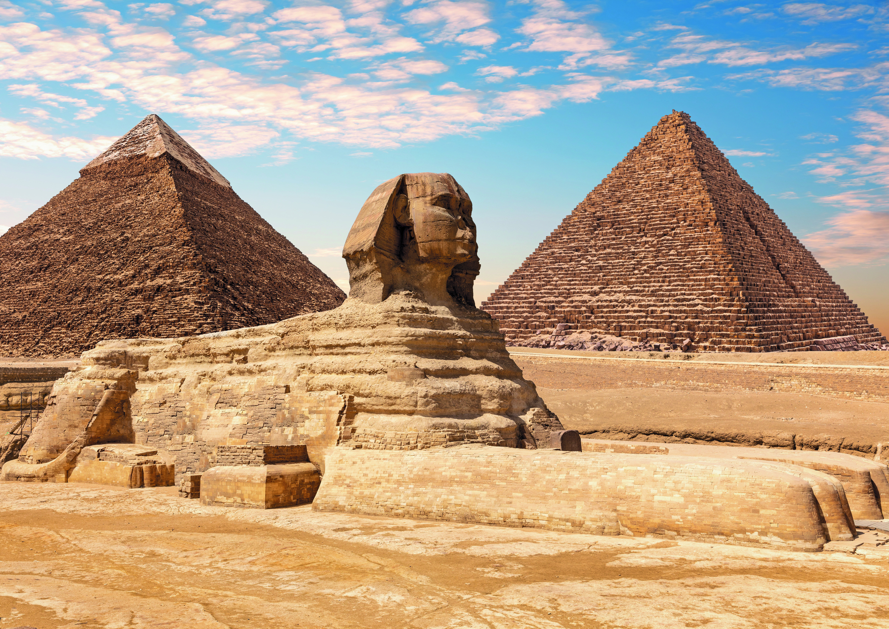 Fotografia. Vista de uma esfinge com rosto humano e corpo de animal, com as quatro patas sobre o solo. Ao fundo, duas pirâmides.