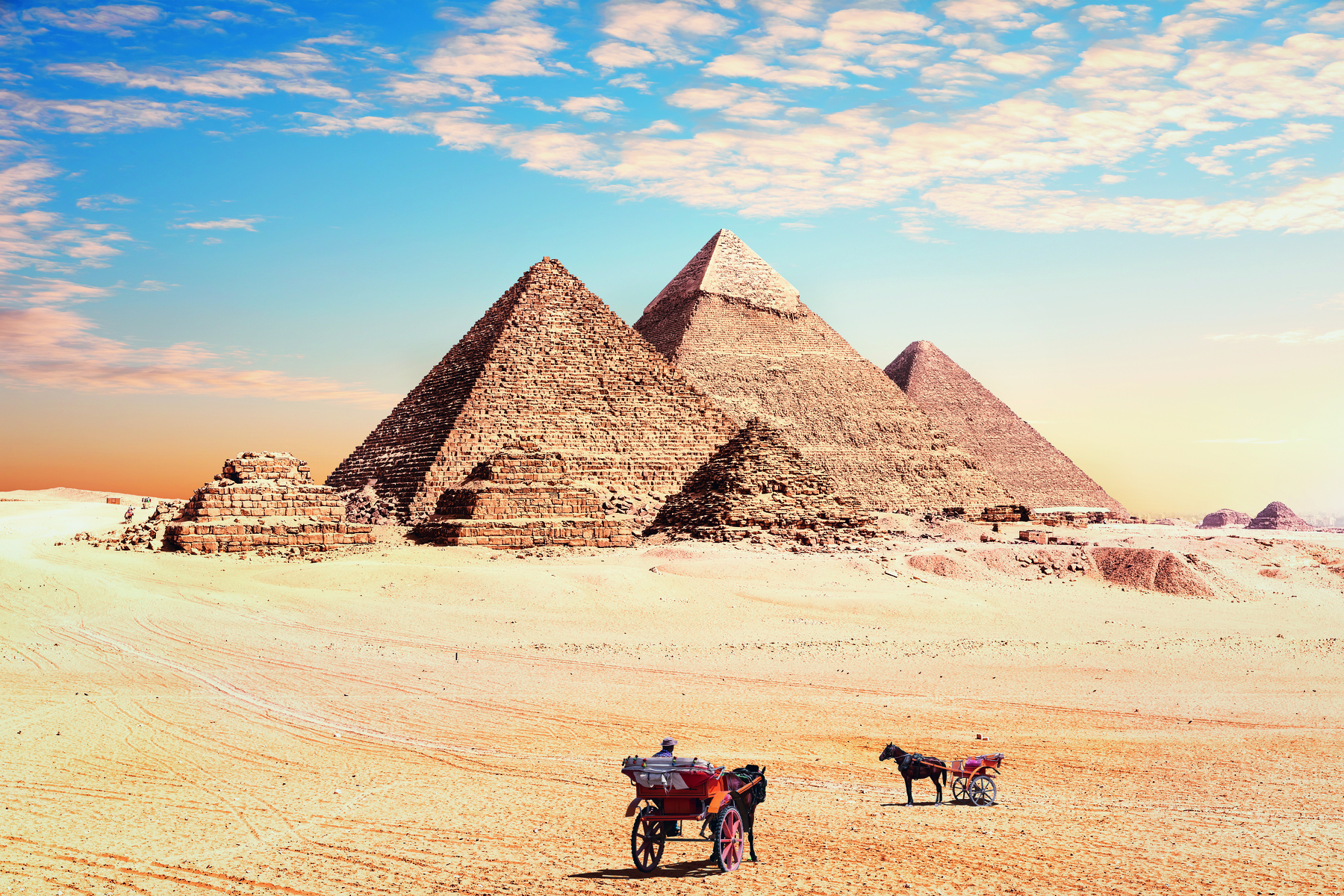 Fotografia. Vista de três grandes pirâmides e três pirâmides menores, com bases quadradas e ápice triangular, sobre um solo arenoso. À frente, duas carroças guiadas por cavalos.