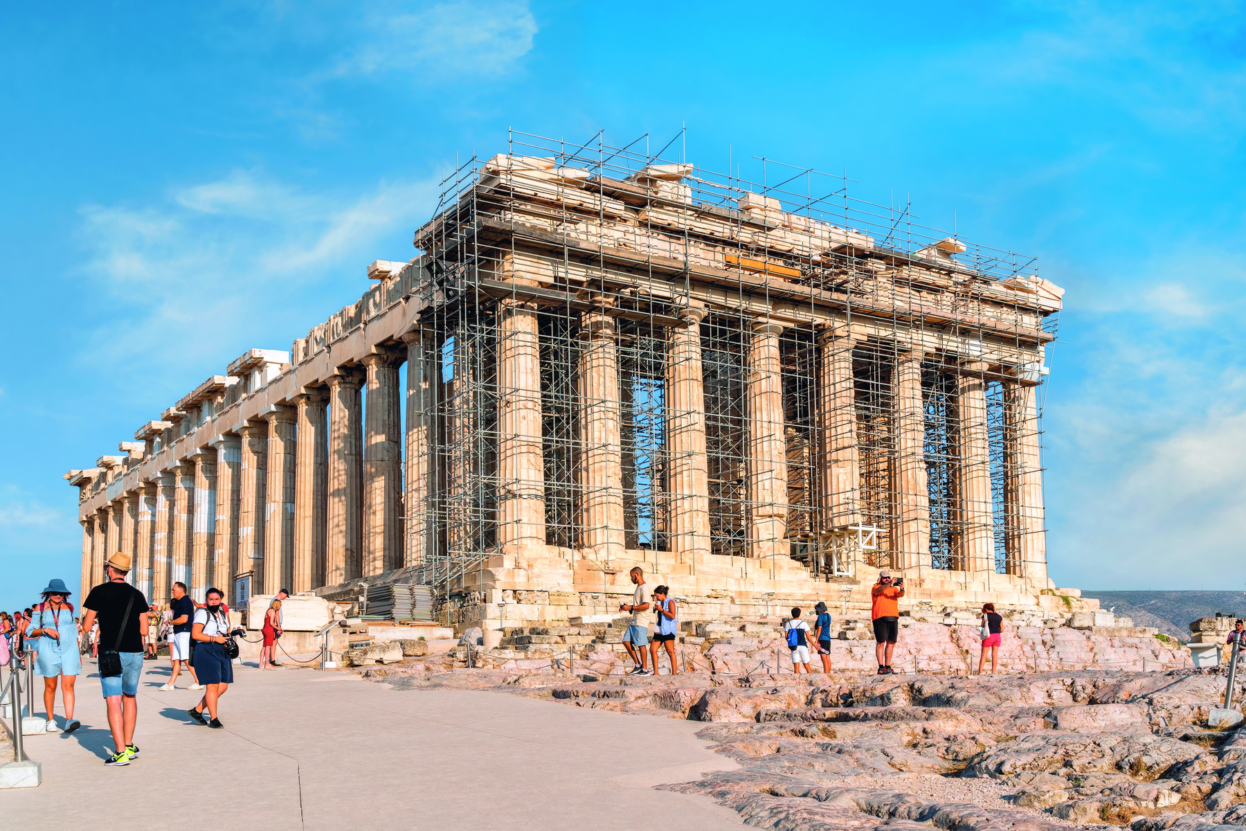 Fotografia. Vista de um templo grego com colunas circulares por todo o perímetro da construção. Ao redor, ruínas de pedras e pessoas transitando.