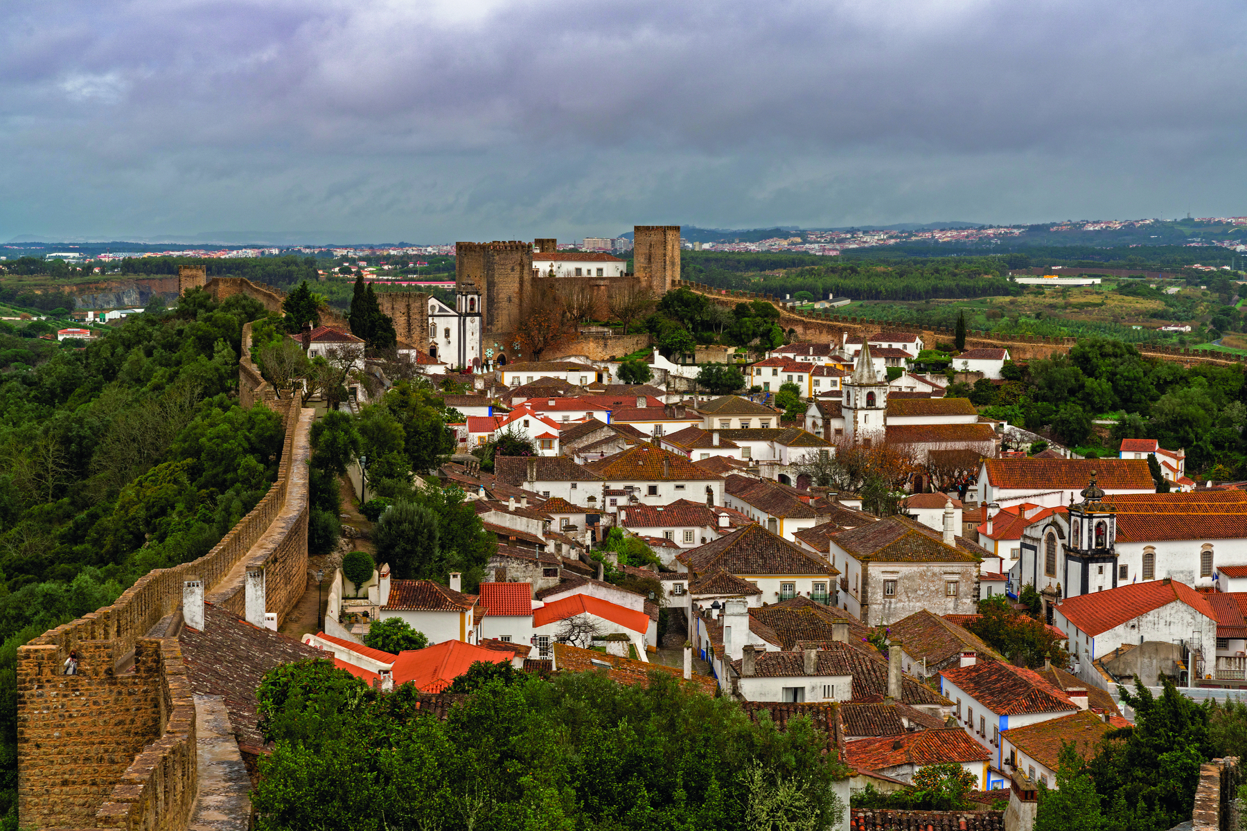Fotografia. Vista aérea de uma cidade com casas brancas e uma extensa muralha de pedras nos arredores. No lado externo da muralha há vegetação densa.