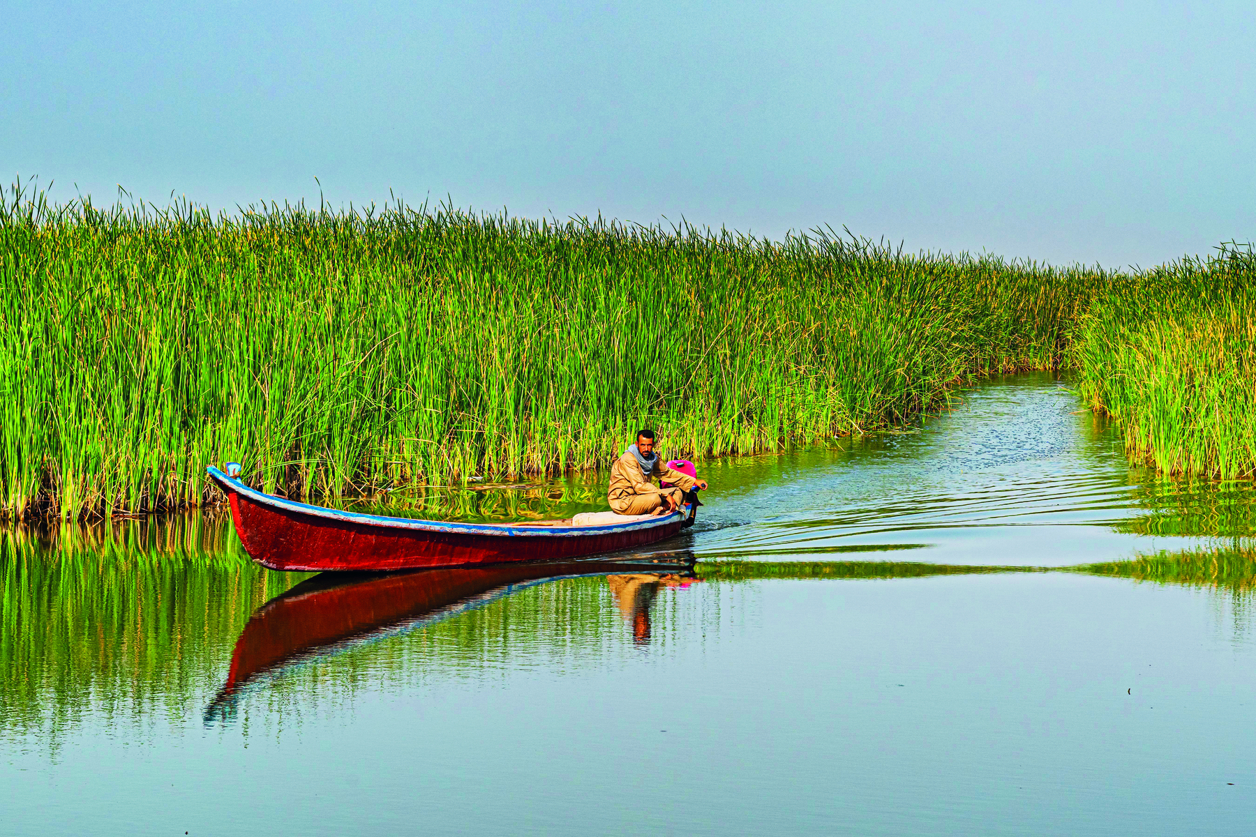 Fotografia. Vista de um homem sentado sobre uma canoa vermelha flutuando em um rio cercado por vegetação densa.