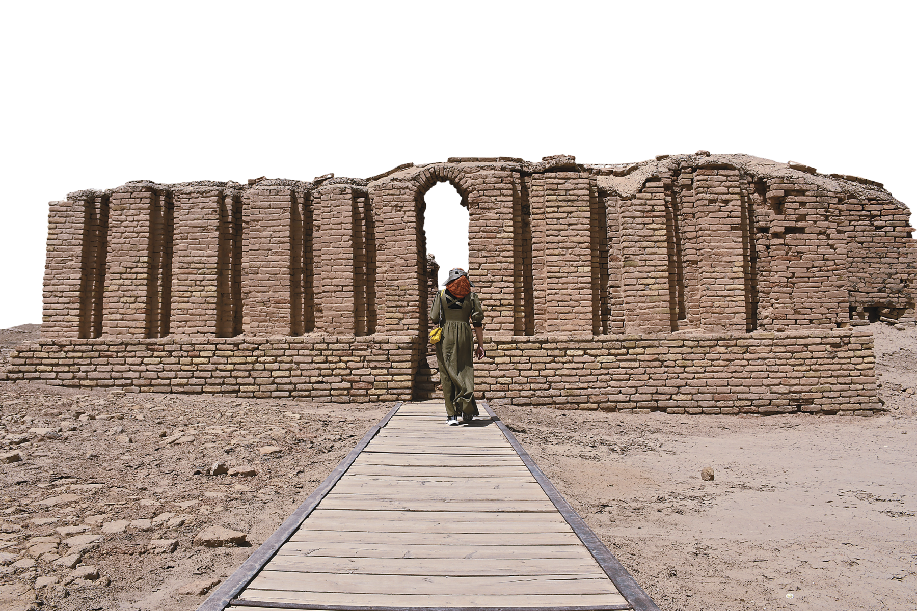 Fotografia. Vista de um muro de tijolos, com pequena abertura central, em ruínas. No centro, uma pessoa em pé sobre um calçamento de madeira rodeado de solo terroso, caminha em direção ao muro.