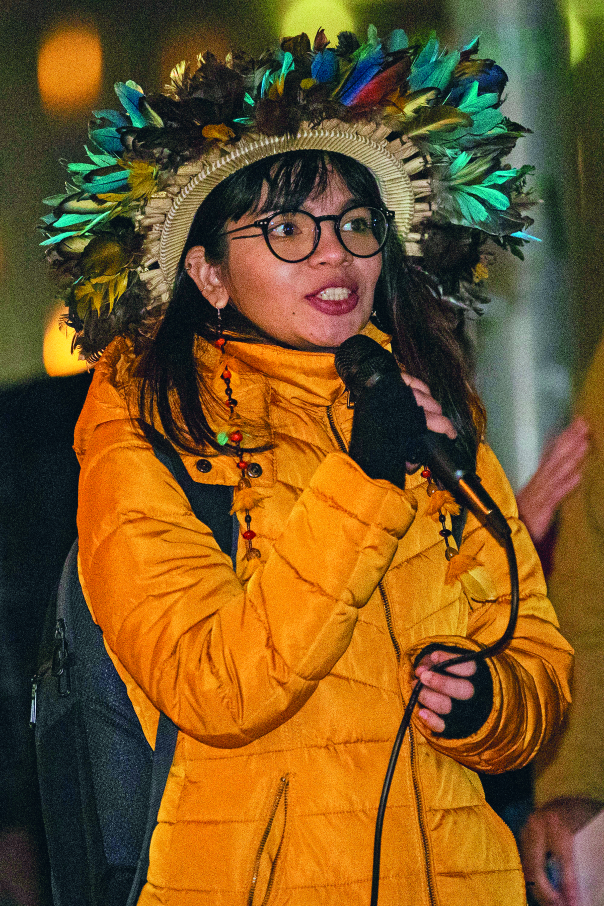 Fotografia. Destaque para mulher indígena, de cabelos longos, lisos e escuros, vestindo uma jaqueta amarela, um par de óculos no rosto e um cocar com penas coloridas na cabeça. Ela segura um microfone com uma das mãos, em direção a sua boca.