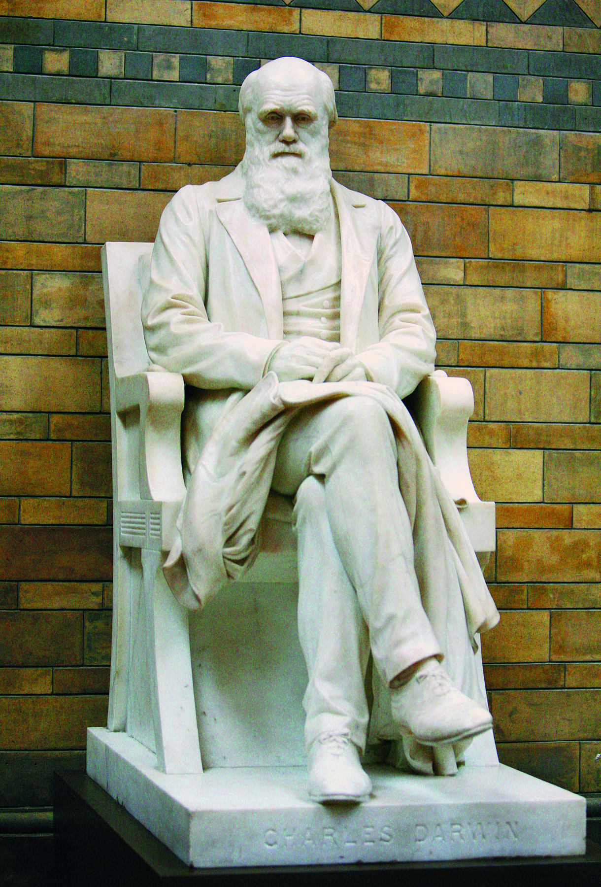 Fotografia. Destaque para escultura em pedra branca, de um homem calvo e barba volumosa, sentado sobre uma cadeira, com as pernas cruzadas e apoiando as mãos sobre o seu colo.