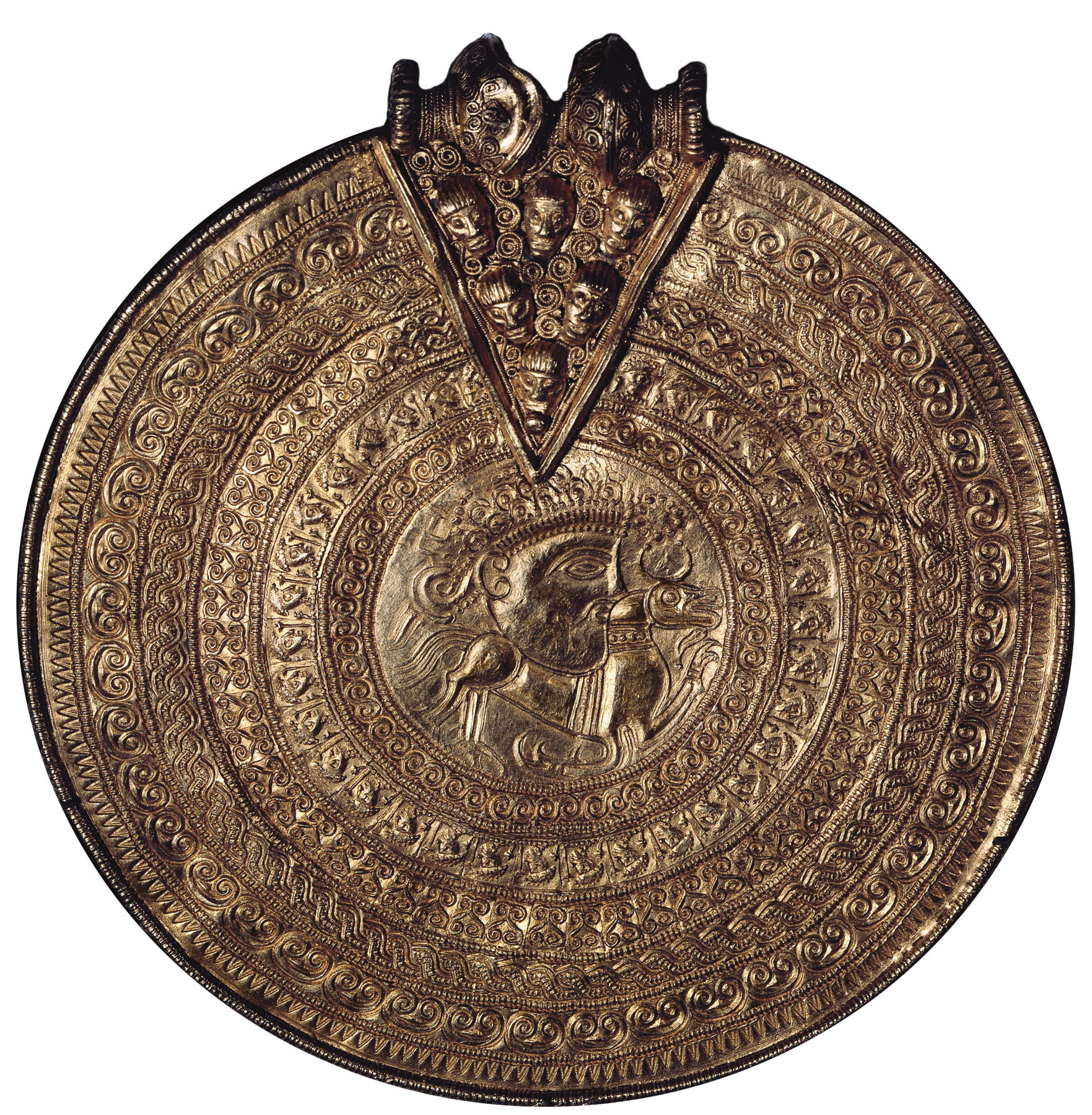 Amuleto. Objeto dourado de formato circular, contendo símbolos ao redor de um rosto visto de perfil ao centro. No topo, há um triângulo entalhado, contendo representações de rostos.