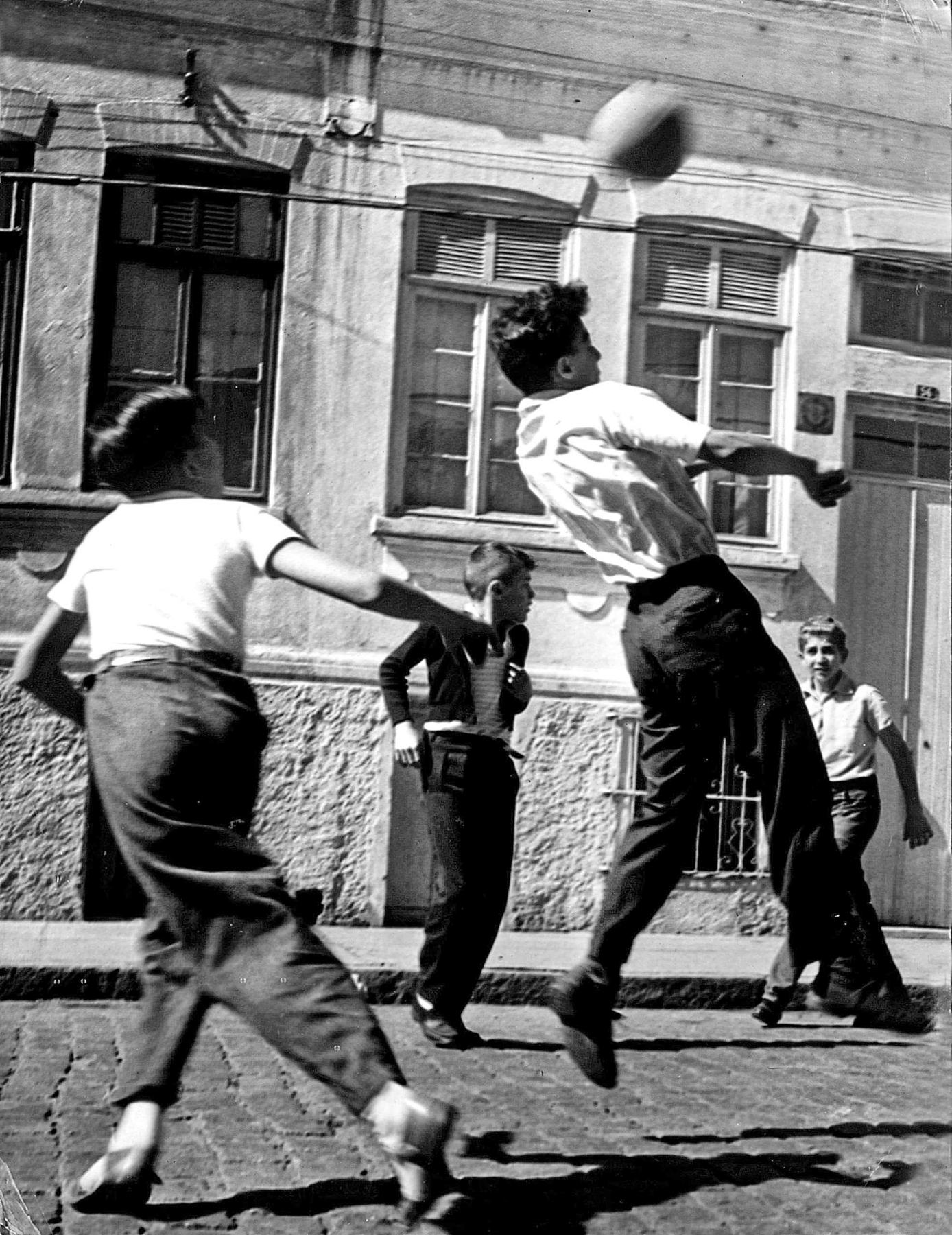 Fotografia em preto e branco. Um grupo de meninos em uma rua com calçamento de pedra, direcionados a uma bola de futebol. Ao fundo, casas.