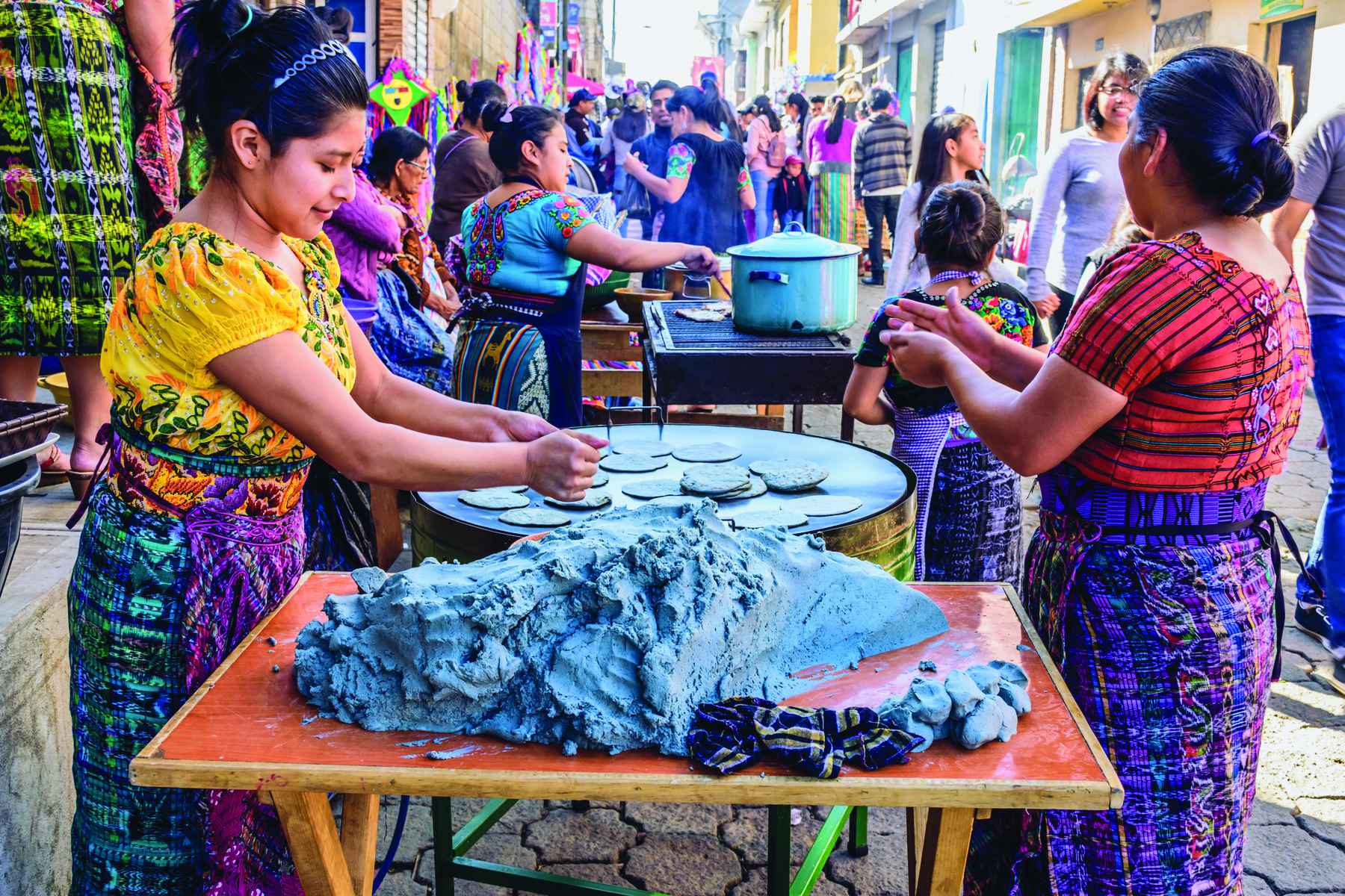 Fotografia. Uma rua de comércio popular abarrotada de pessoas. Destaque para duas mulheres preparando massas finas e arredondadas, com o auxílio das mãos.