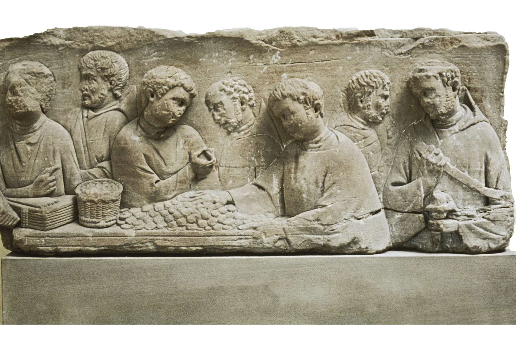Relevo. Imagem entalhada em pedra de homens vestindo túnicas, lado a lado, próximos a uma mesa contendo cestos e muitas moedas.