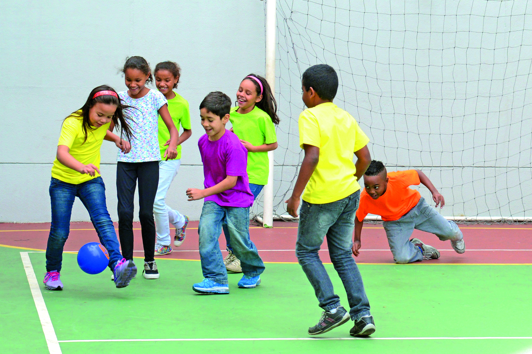 Fotografia. Um grupo de meninas e meninos em uma quadra esportiva, direcionadas a uma bola de futebol. Ao fundo, uma trave branca retangular e uma rede trançada.