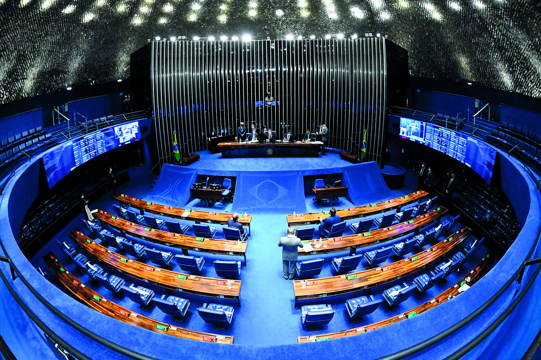 Fotografia. Vista superior de um palanque com uma mesa central, imagem da bandeira do Brasil, e à frente, uma sequência de mesas e cadeiras enfileiradas. A área é cercada por paredes circulares azuis.