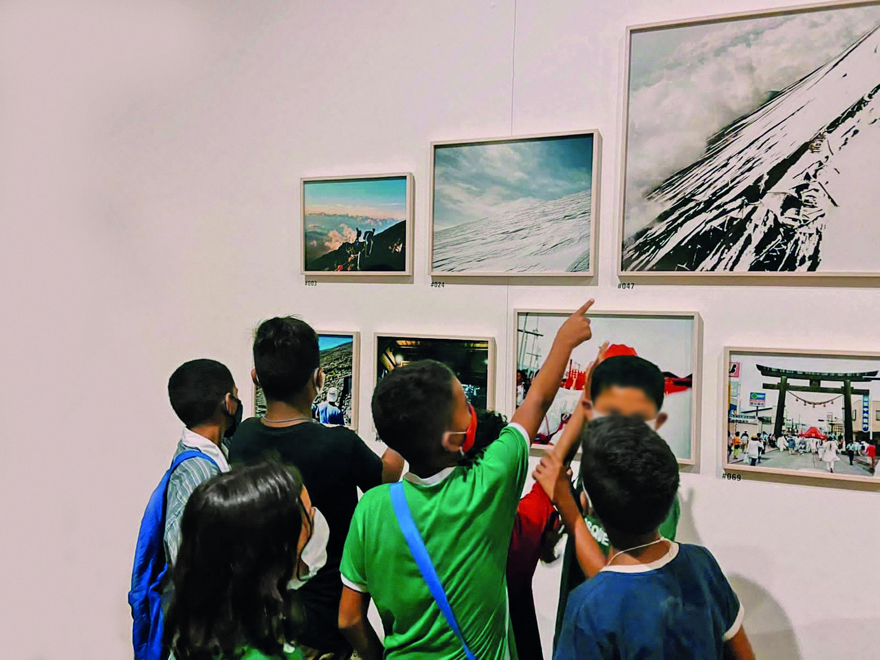 Fotografia. Um grupo de crianças a frente de quadros em exposição sobre uma parede branca.