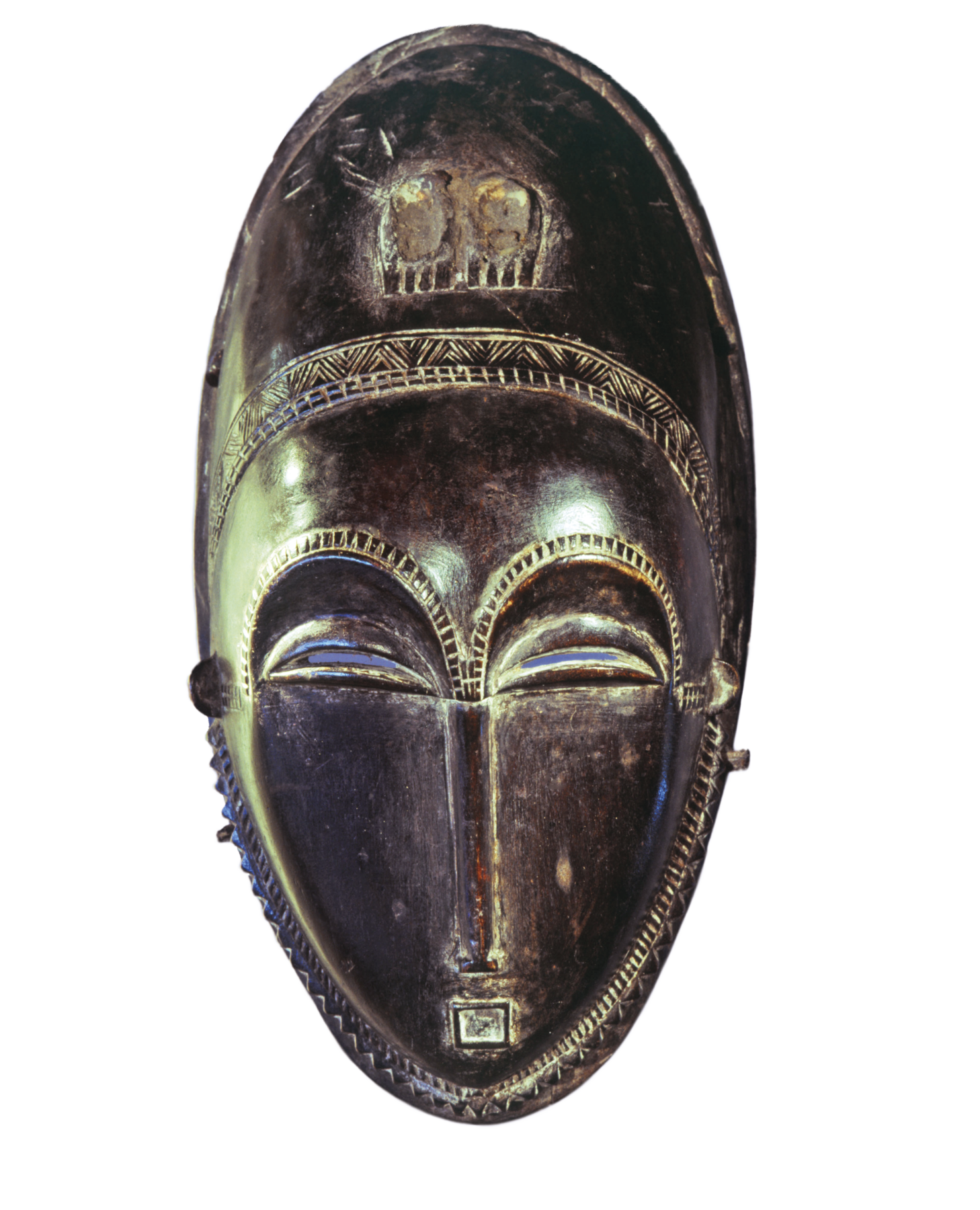 Máscara. Objeto de madeira, em formato oval, com a representação de olhos semifechados, nariz afilado e boca pequena, contornado por entalhes lineares.