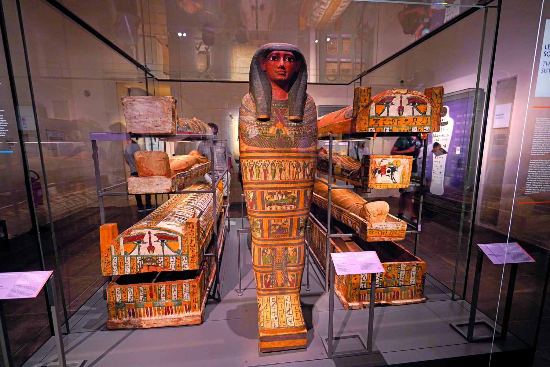 Fotografia. Exposição de vários sarcófagos egípcios em posição horizontal, com destaque para um, ao centro, em posição vertical, apresentando a escultura de um rosto humano no topo, com longos cabelos escuros, e ilustrações por toda a superfície.