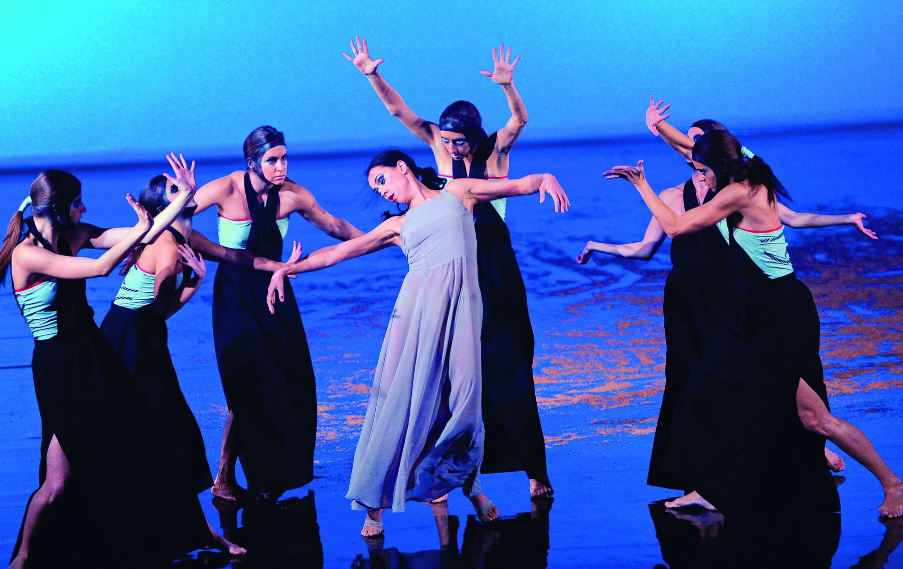 Fotografia. Imagem de uma apresentação em um palco, com mulheres em posições de danças, com os braços levantados em diferentes ângulos. Ao centro, destaque para uma mulher de vestido azul claro, cercada de outras mulheres de roupas escuras com faixas azuis.