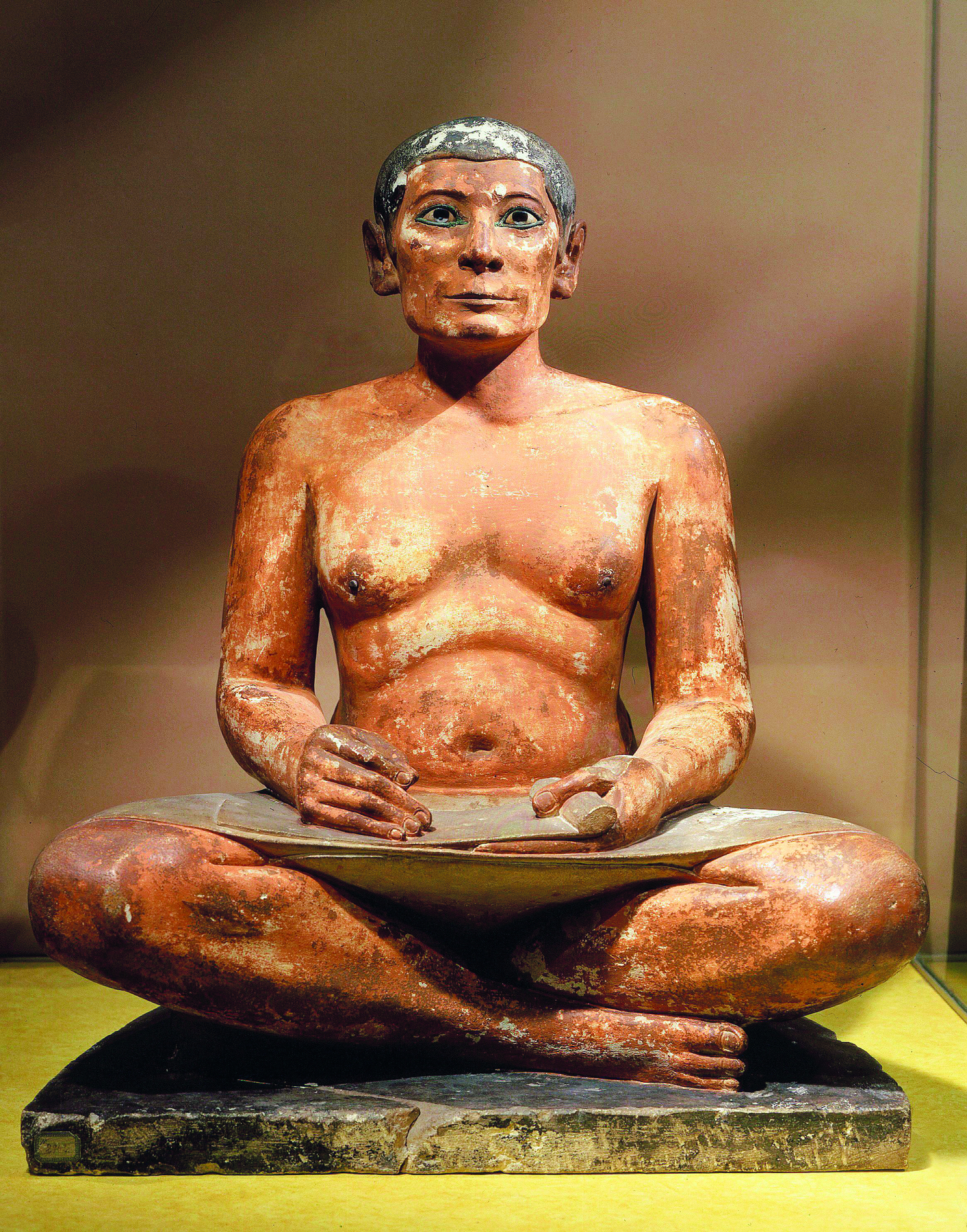 Escultura. Um homem com peito desnudo, sentado no solo com as pernas cruzadas, apoiando uma folha de papiro sobre seu colo.