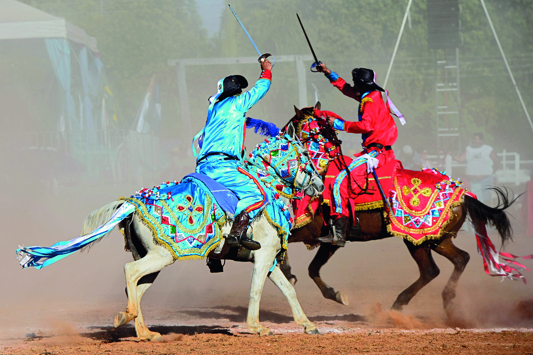 Fotografia. Dois homens montando cavalos, segurando espadas esticadas para cima. O homem da esquerda veste camisa e calça azuis, o homem da direita veste camisa e calça vermelhas. Os cavalos tem o dorso coberto por capas na mesma cor do traje de seus cavaleiros.