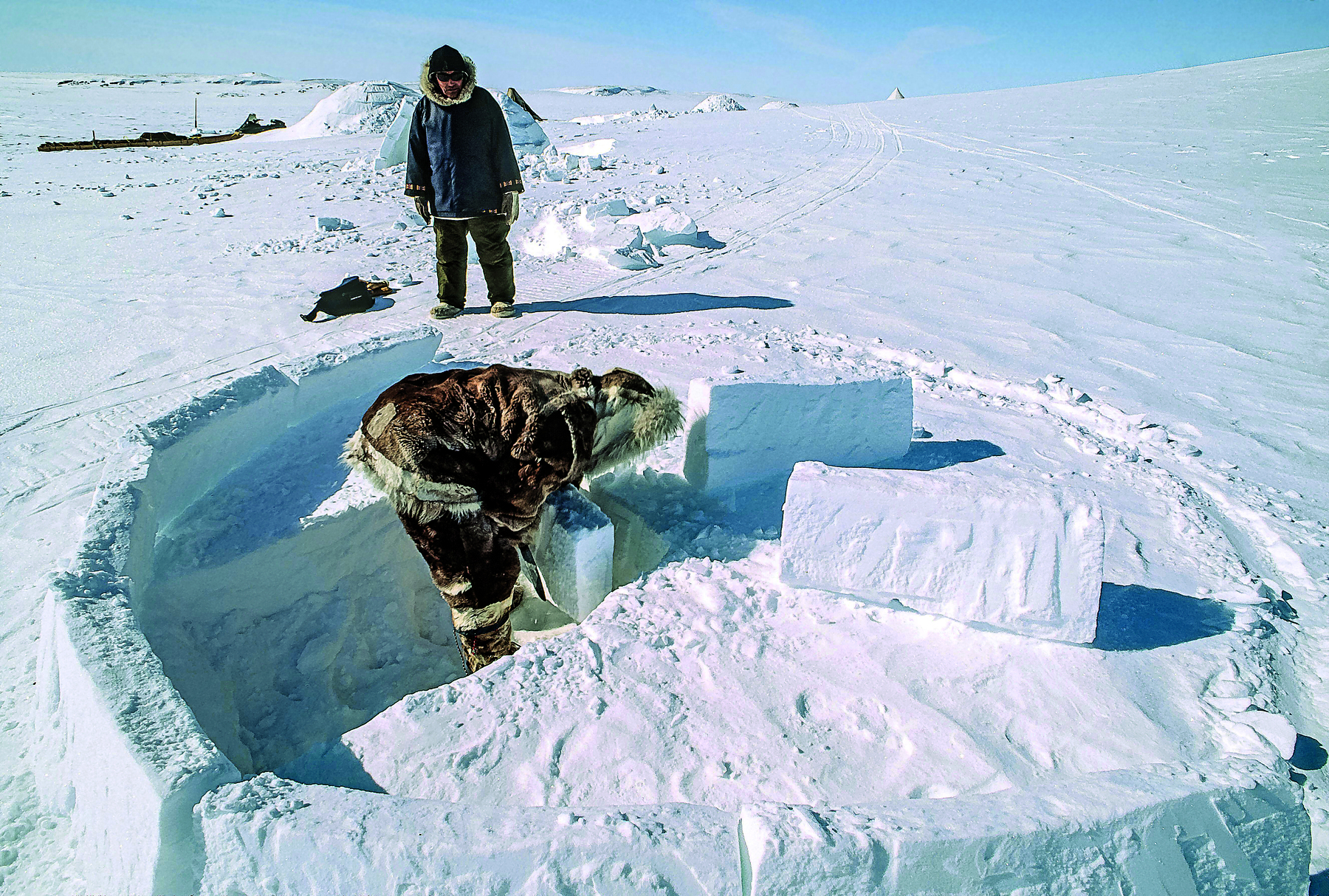 Fotografia. Destaque para dois homens vestindo roupas grossas com pelos nas extremidades e capuzes ao redor da cabeça. Ambos estão em um terreno completamente coberto de neve, próximos a blocos de gelo, organizados em círculo.
