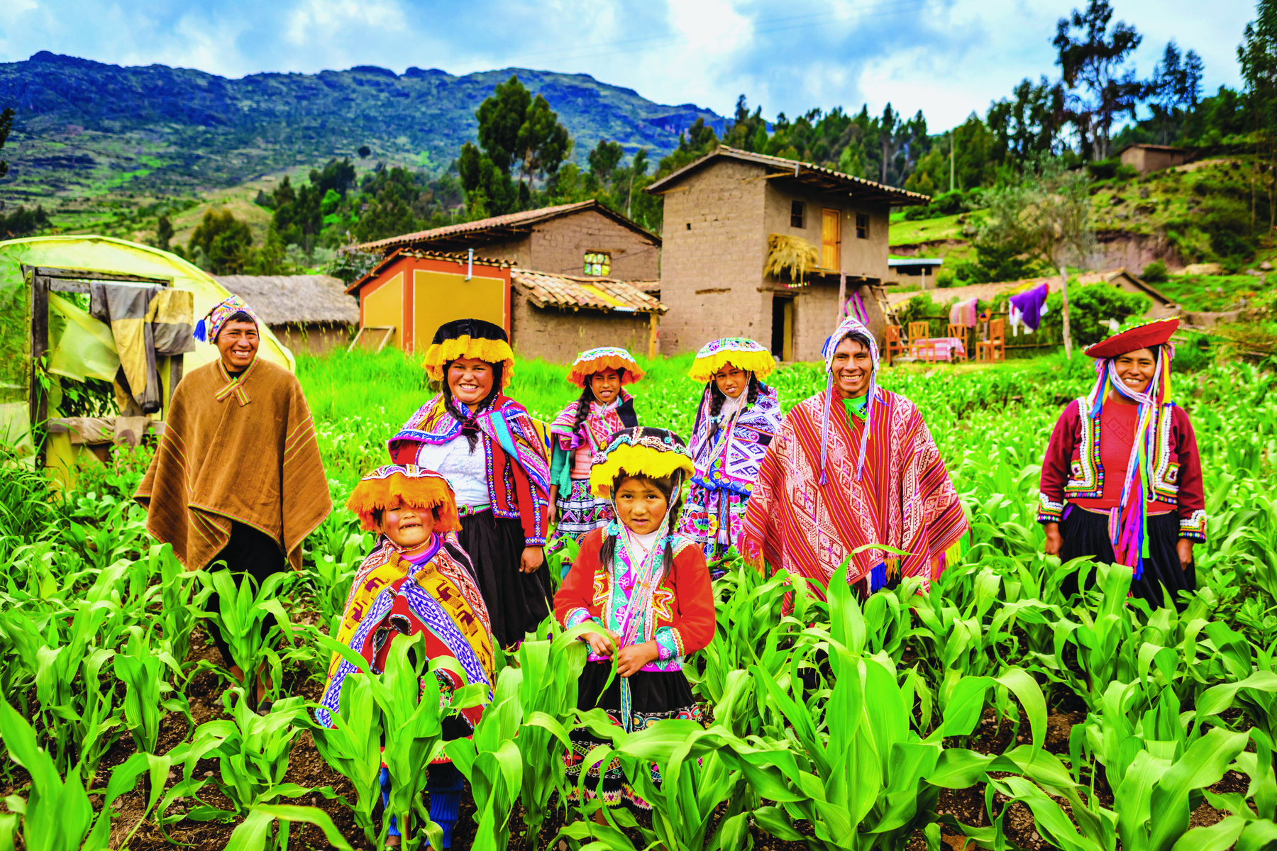 Fotografia. Um conjunto de adultos e crianças no meio de uma plantação, todos vestindo túnicas e capas coloridas e chapéus largos. Em segundo plano há casas de tijolos com tetos triangulares.