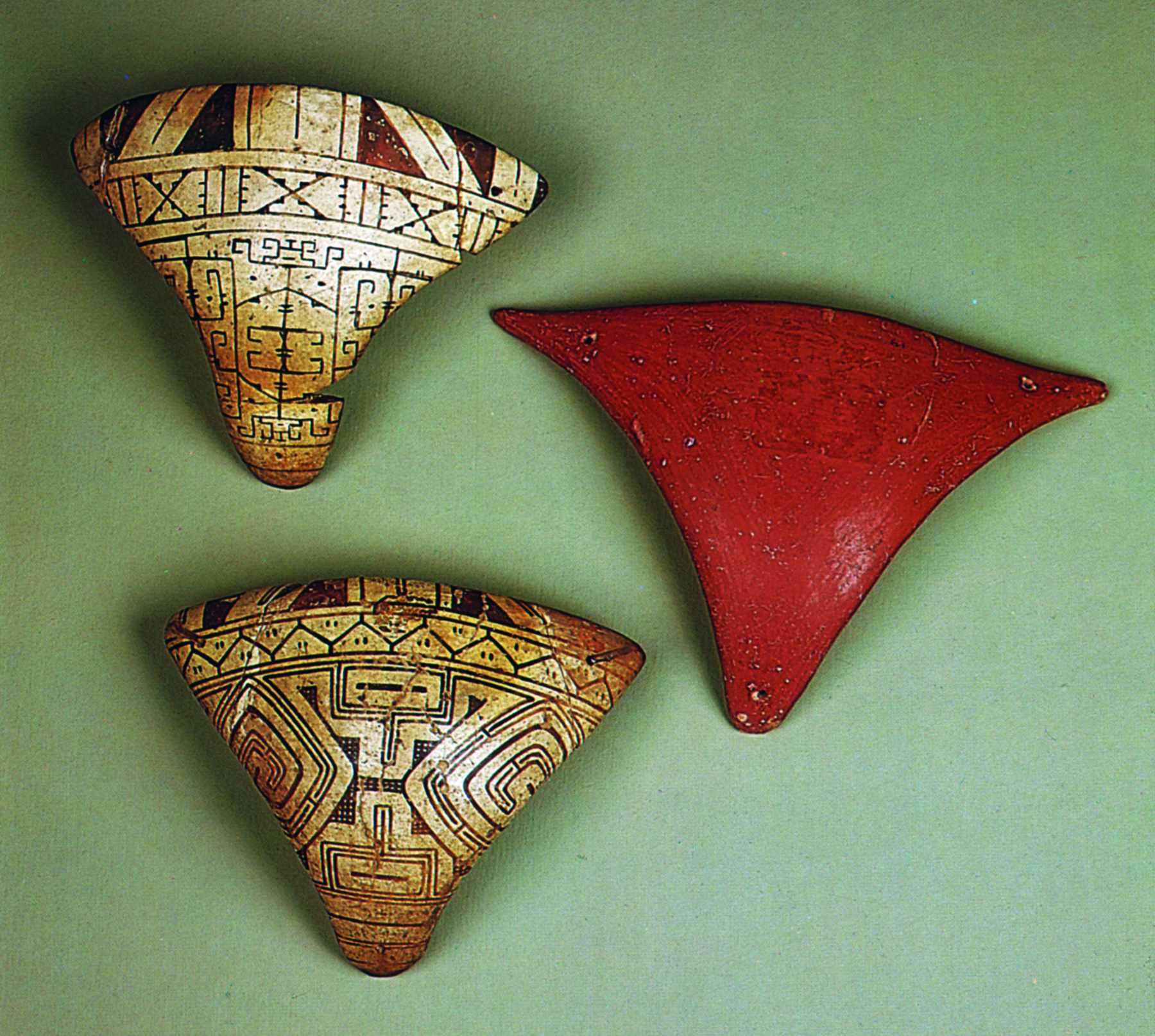 Cerâmica. Três objetos triangulares, levemente curvos. Dois deles possuem desenhos de linhas e formas geométrica. O outro é liso, com superfície avermelhada.