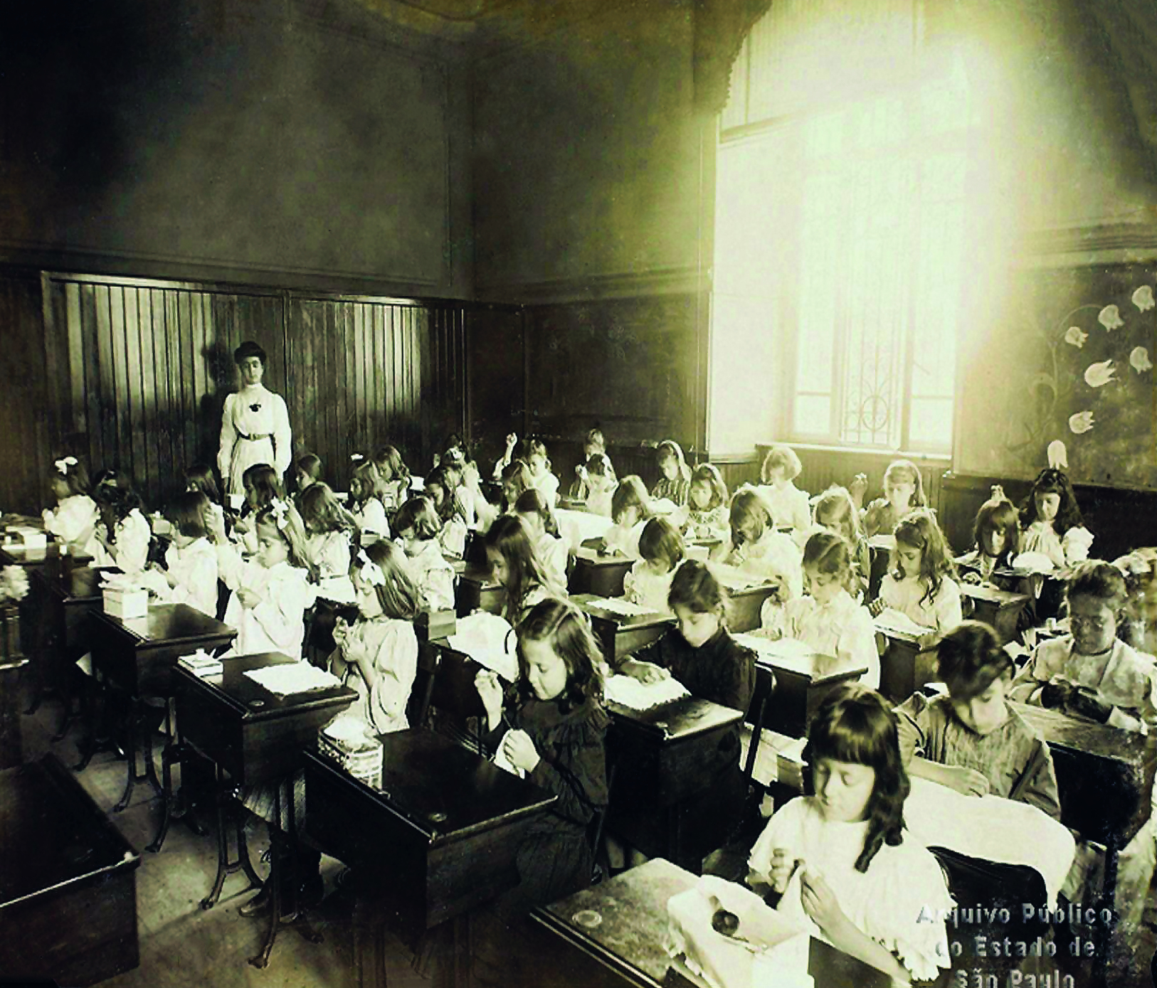 Fotografia em preto e branco. Uma sala de aula formada por alunas uniformizadas, sentadas sobre carteiras escolares de madeira. Ao fundo, uma mulher adulta, em pé.
