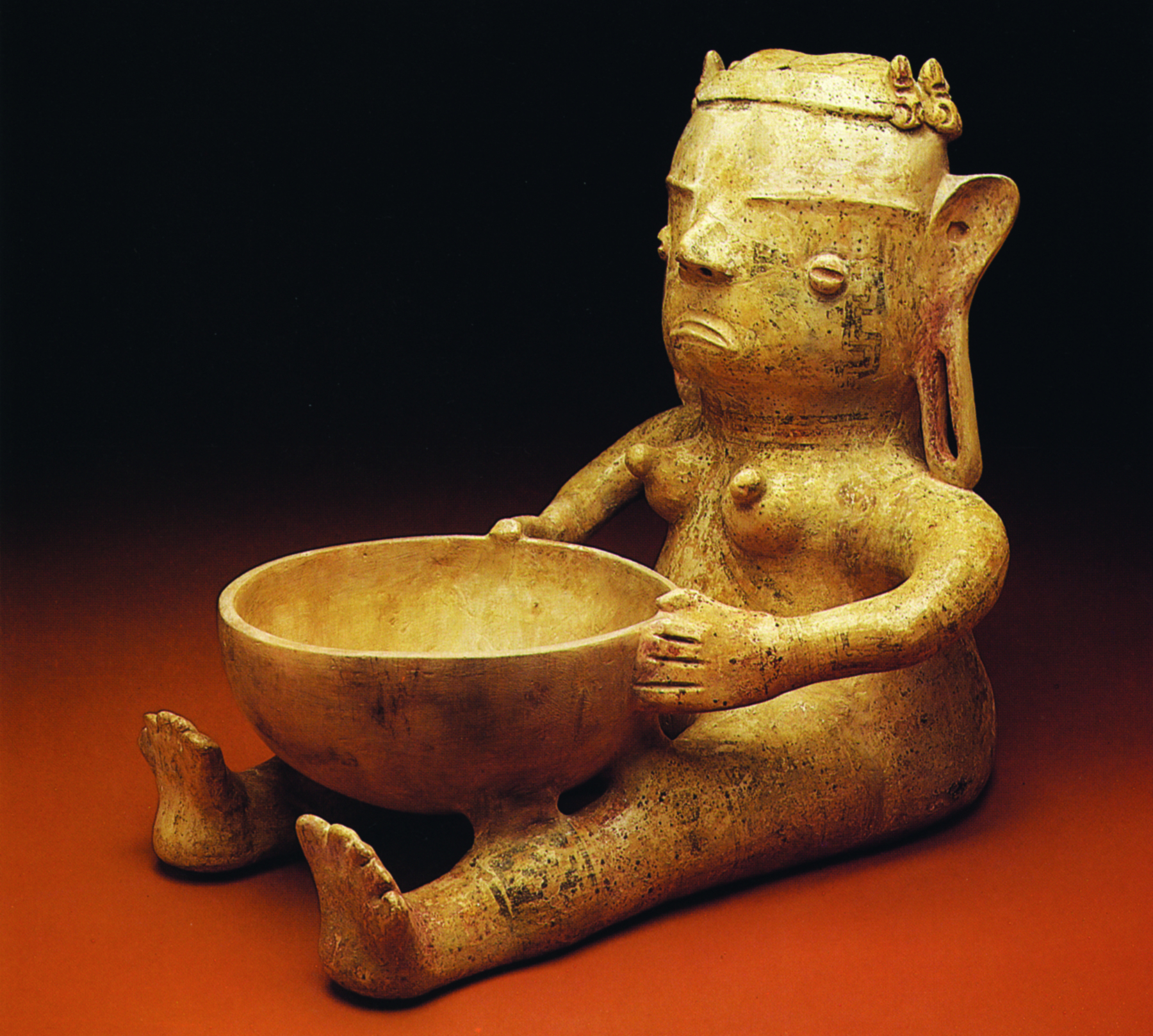 Cerâmica. Escultura de uma figura feminina sentada, com as pernas esticadas e as mãos nas laterais de um recipiente arredondado, apoiado sobre suas pernas.