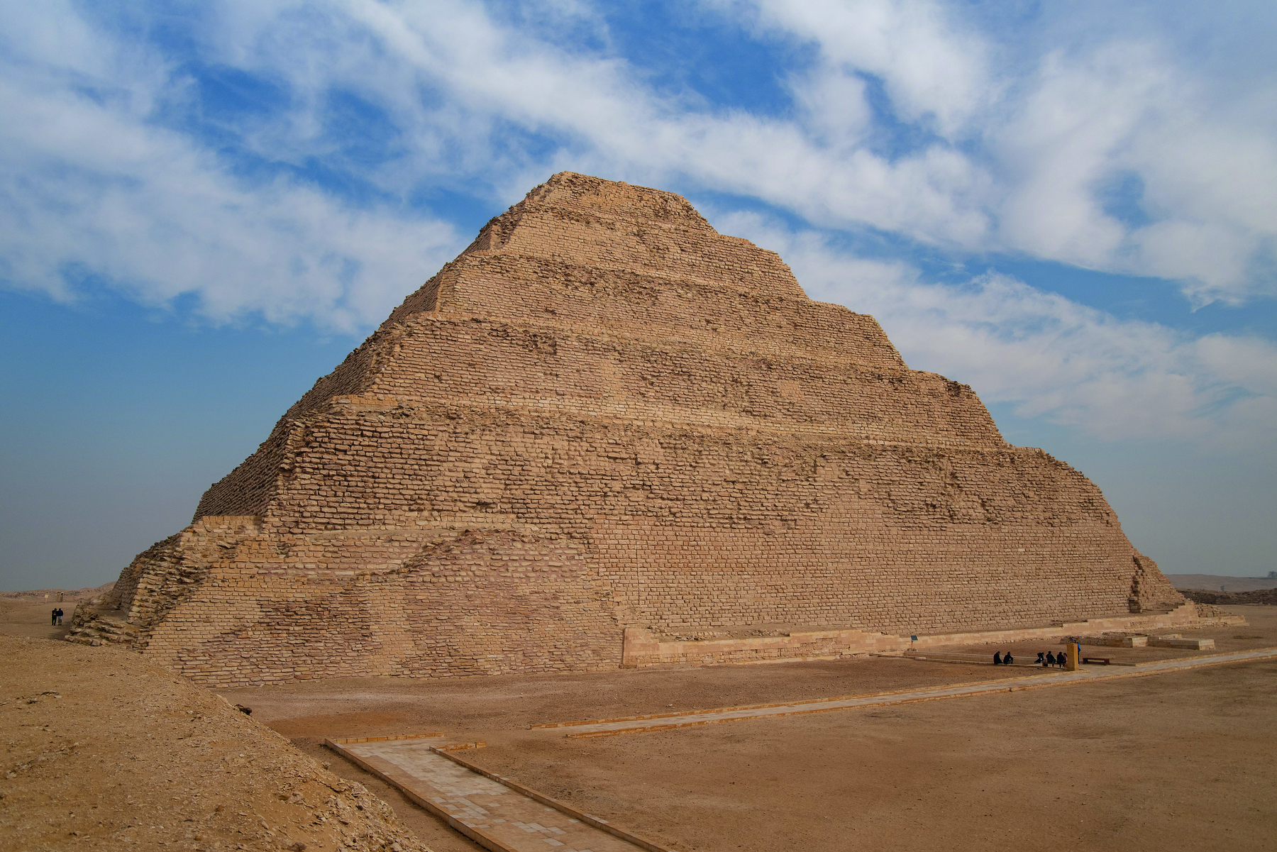 Fotografia. Vista de uma pirâmide em níveis retangulares, decrescentes da base para o ápice. Ao redor, terreno arenoso e céu azul com nuvens.