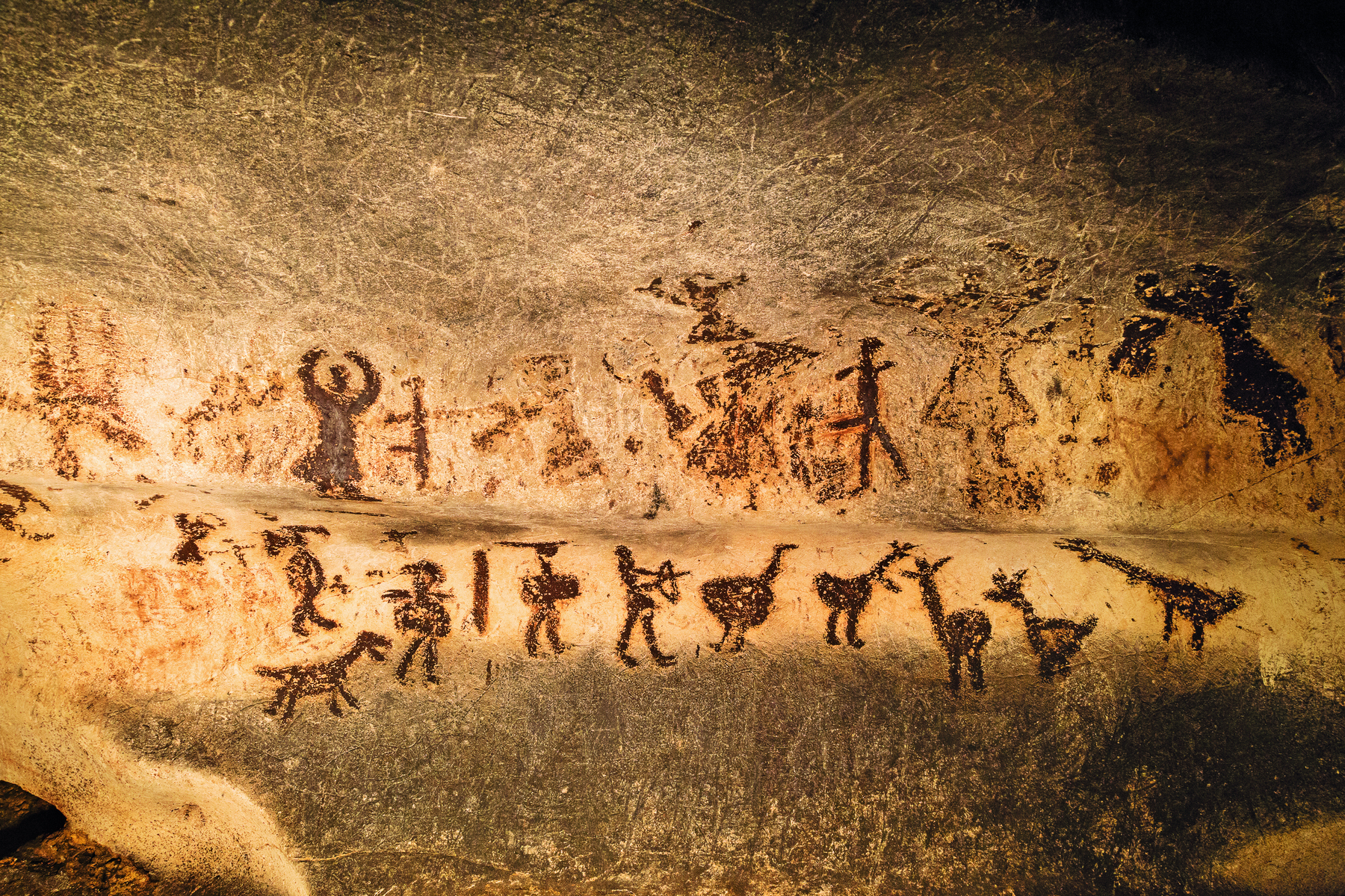 Fotografia. Parede de uma caverna contendo pinturas rupestres, representando animais e pessoas portando diferentes instrumentos, como lanças  e arcos.