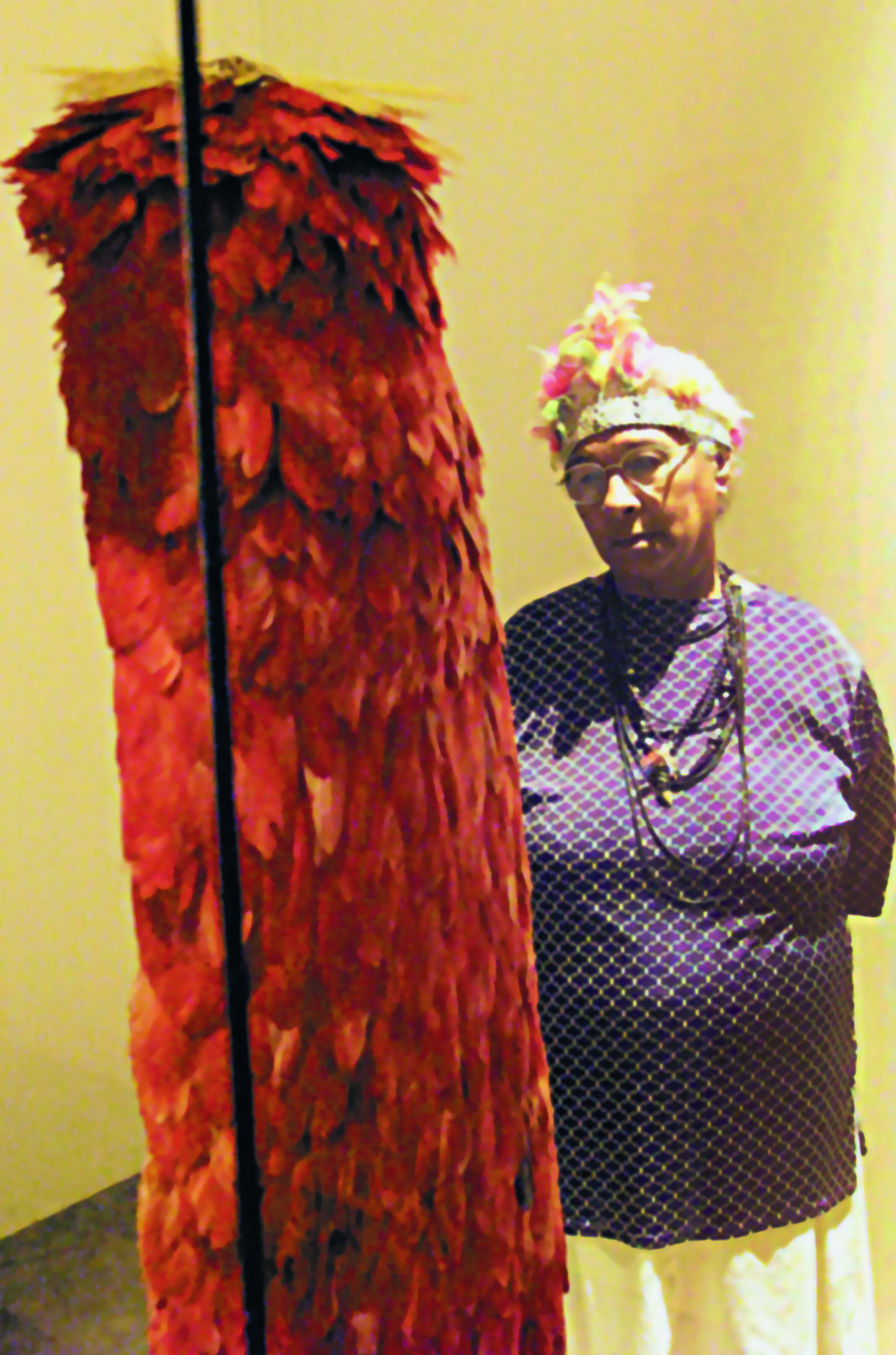 Fotografia. Uma mulher indígena de cabelos grisalhos, curtos e lisos, vestindo uma camisa azul e saia branca, um cocar com penas e colares ao redor do pescoço, observa um manto comprido coberto de penas vermelhas.