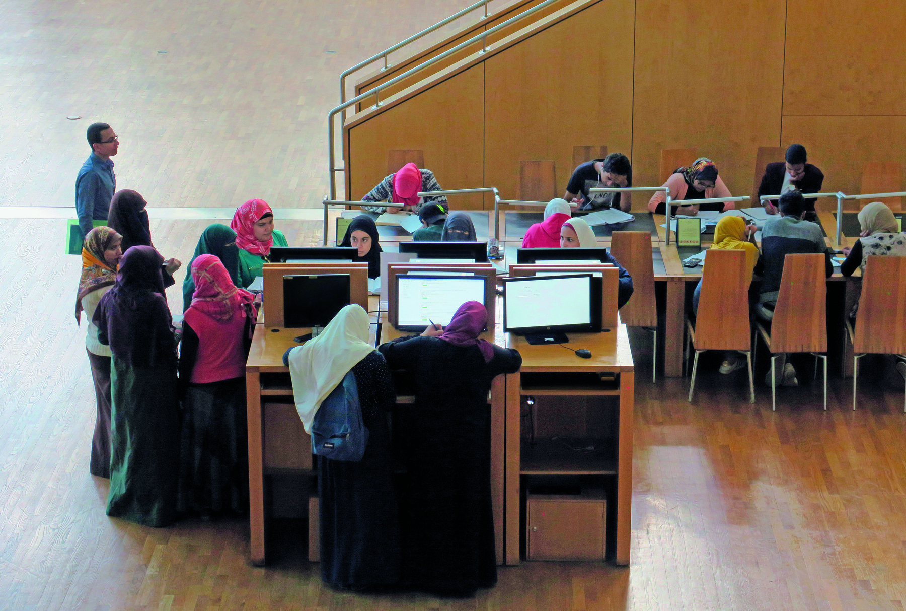 Fotografia. Imagem de estudantes no interior de uma biblioteca, próximas à telas de computadores sobre suportes de madeira. As mulheres utilizam véus de diferentes cores e estampas, que cobrem a cabeça e o pescoço.