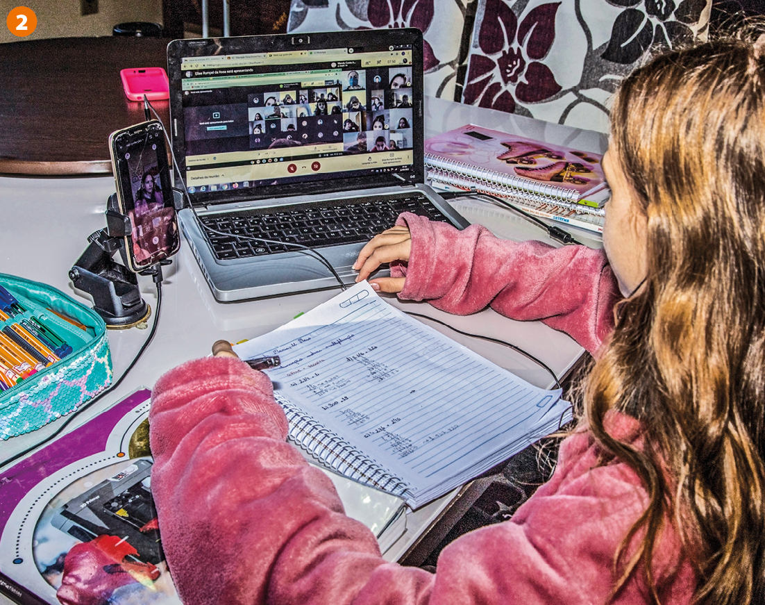 Fotografia. Uma menina de cabelos loiros, longos e lisos, vista de perfil, vestindo uma jaqueta rosa, sentada a frente de uma mesa contendo caderno, livro, estojo com canetas, um aparelho de celular e um computador ligado. Em seu caderno há contas de matemática.