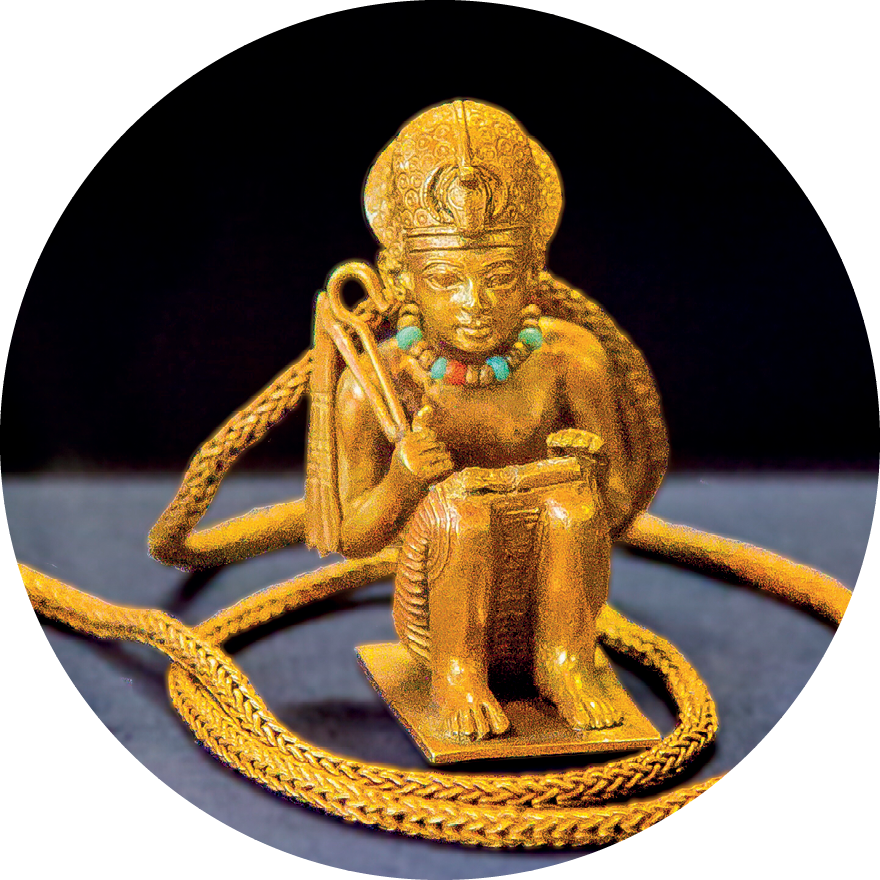 Estatueta. Objeto dourado representando um homem sentado no solo, com os joelhos flexionados para frente, segurando hastes em uma de suas mãos. Apresenta colar ao redor de seu pescoço e uma coroa adornada na cabeça.