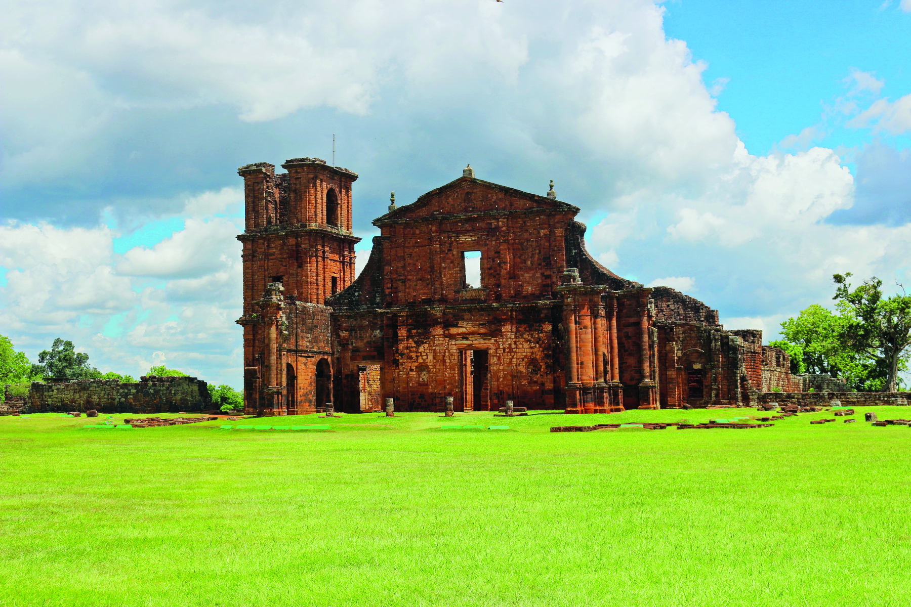 Fotografia. Vista da fachada de uma igreja em ruínas sobre um campo gramado, com paredes feitas em tijolos, uma torre lateral à esquerda, e uma porta central retangular.