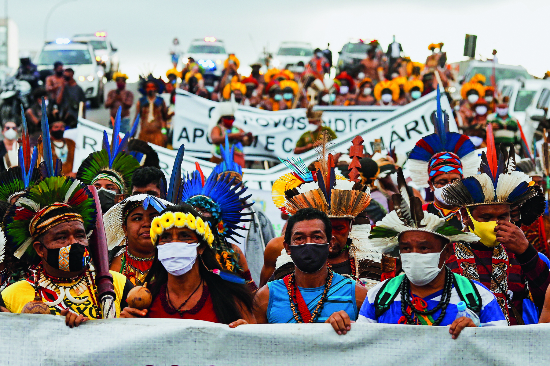 Fotografia. Uma multidão de homens e mulheres indígenas, utilizando cocares com penas coloridas e colares sobre seus peitos, lado a lado, segurando faixas com textos.