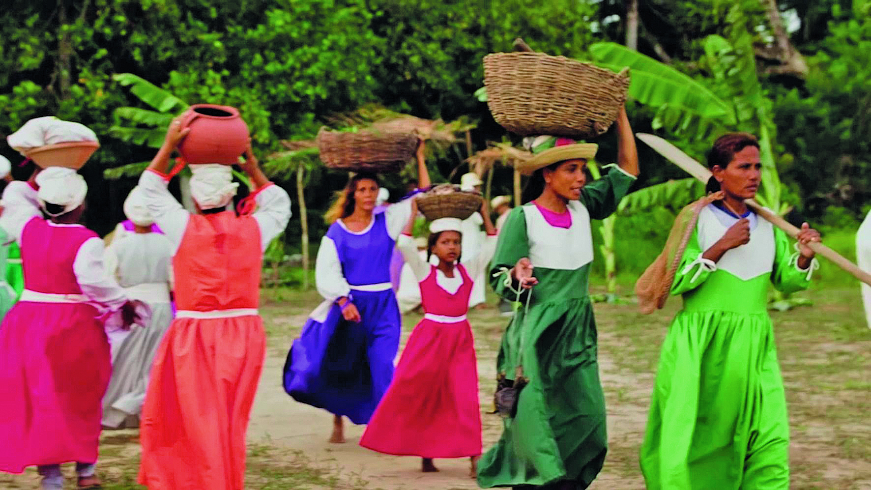 Fotografia. Um conjunto de mulheres em pé sobre um campo gramado, próximas umas às outras, cada uma delas utilizando um vestido longo com cores diferentes, apoiando objetos sobre suas cabeças, como cestas, bacias e tigelas. Ao fundo, árvores e plantas.