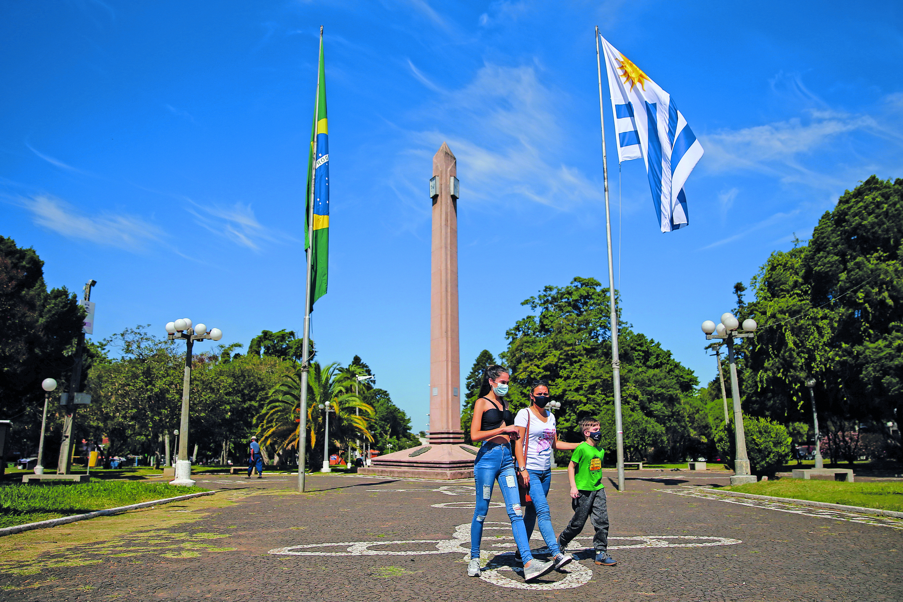 Fotografia. Uma jovem, uma mulher e uma criança, em pé sobre uma praça, à frente de um monumento longo e vertical, cujas laterais apresentam duas hastes com as bandeiras do Brasil e Uruguai. Às laterais, áreas arborizadas.