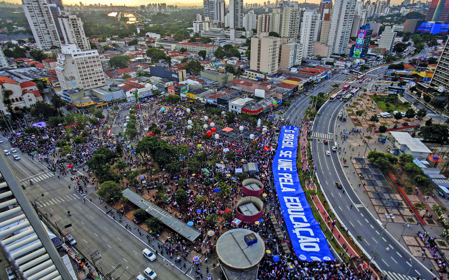 Fotografia. Vista aérea de uma área urbana contendo uma manifestação de pessoas cercadas por avenidas e edifícios. Há uma longa faixa azul estendida sobre os manifestantes, com o texto: 'O Brasil se une pela Educação'.