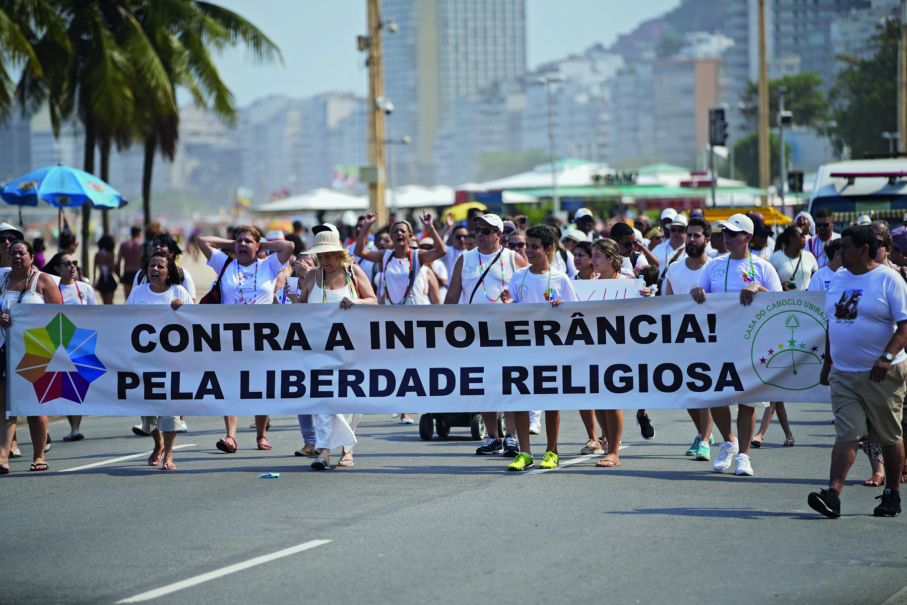 Fotografia. Uma multidão de pessoas sobre uma via pavimentada, todos vestindo camisetas brancas, segurando uma larga faixa com o texto: 'Contra a intolerância! Pela liberdade religiosa'.
