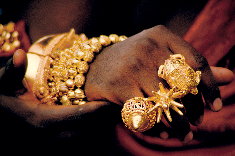 Fotografia. Destaque para a mão de uma pessoa com anéis e braceletes dourados. Os anéis, possuem formatos de conchas e de tartaruga. Os braceletes contornam o pulso.
