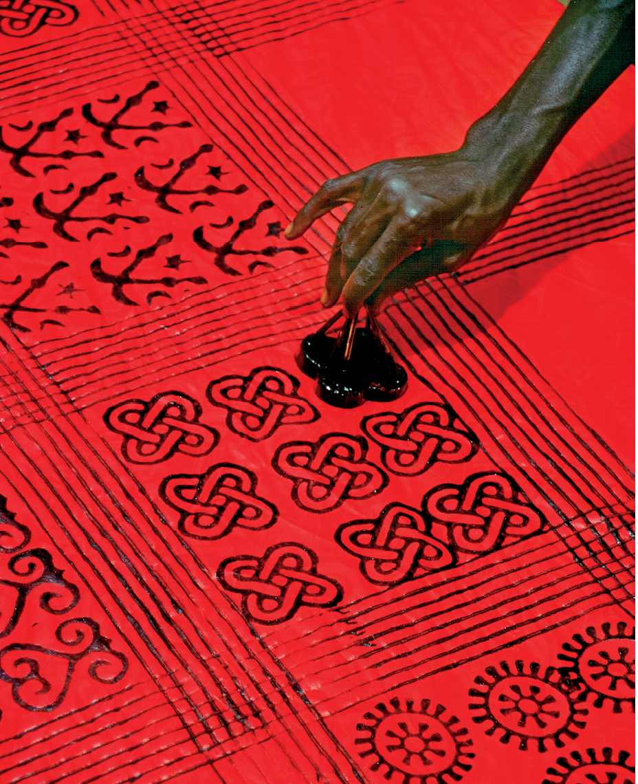 Fotografia. Destaque para a mão de uma pessoa segurando um pequeno objeto com marcação de linhas entrelaçadas sobre um tecido vermelho estendido com detalhes e contornos pintados em preto.
