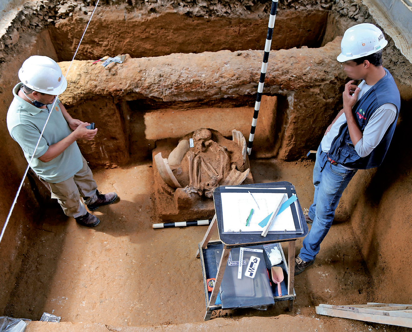 Fotografia. Destaque para dois homens em pé, sobre uma área escavada, com destaque para uma urna contendo uma ossada humana.